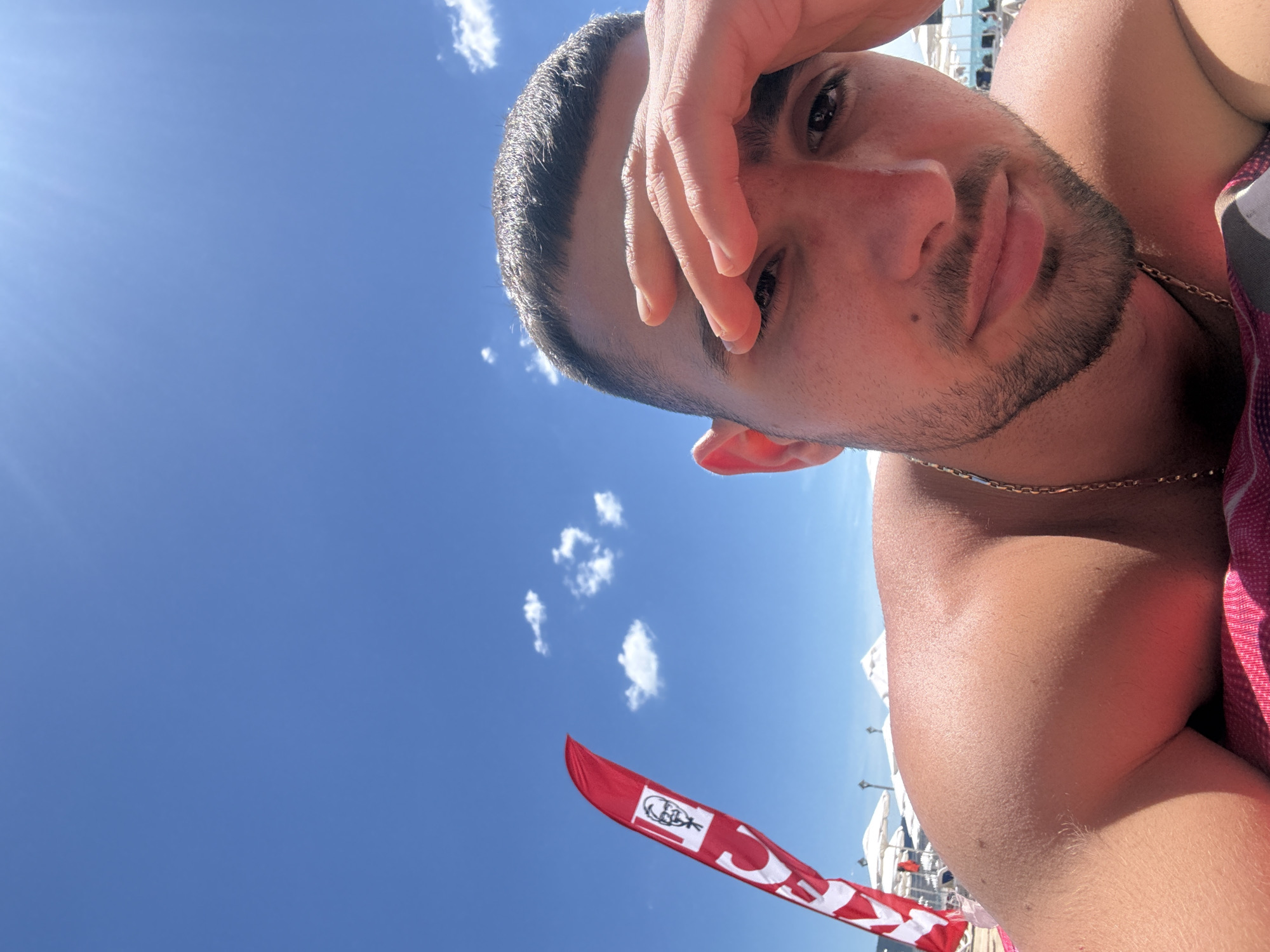 The image shows a man lying on a pink towel on a sunny day at a beach, likely in the city of Nesebar, Bulgaria, given the presence of a red flag with the KFC logo, a common sight at tourist locations. The man is wearing a gold chain and has his hand over his forehead, as if shielding his eyes from the sun. In the background, there are white beach umbrellas and what appears to be the sea. The sky is bright blue with a few scattered clouds. The scene is peaceful and suggests a relaxing day at the beach.