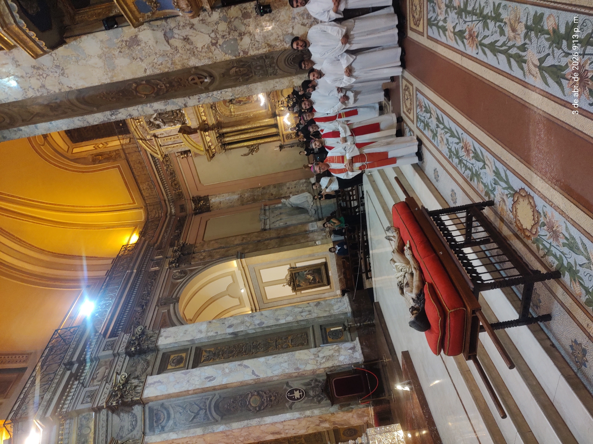 Here is a description of the image:
**Scene Description:**
The image depicts the interior of a large, ornate church. The architectural details include marble columns, arches, and a domed ceiling, which is illuminated by natural light. Intricate patterns and designs are visible on the walls. A group of people, dressed in white robes and red stoles, are standing in a line. A red coffin on a stand is in the foreground. The time stamp indicates "3 de abr. de 2026 9:13 p. m."
**People:**
A group of priests are standing in a line
**Objects:**
Marble columns, arches, a domed ceiling, walls with intricate patterns and designs, red coffin on a stand, white robes and red stoles, church.
**Environment:**
The environment is in the interior of a large, ornate church with natural light.
**Location:**
City: Unknown
Location: Unknown