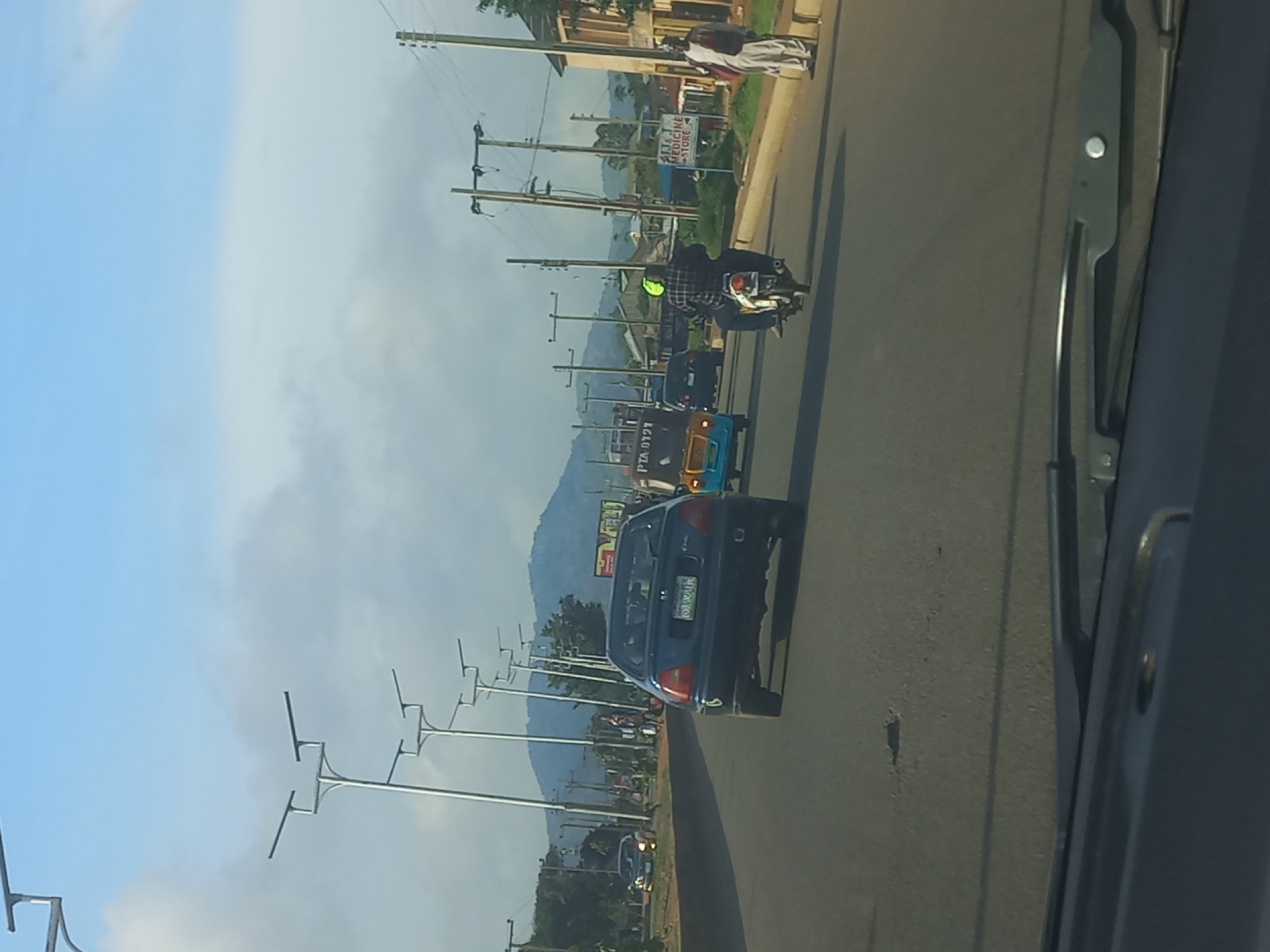 The image shows a street scene in a city, with several vehicles and pedestrians visible. In the foreground, the hood and windshield wipers of a car are visible, suggesting the photo was taken from inside a vehicle. The road appears to be paved and there are streetlights lining the side of the road.
Several vehicles are on the road, including a dark blue sedan, a blue and yellow auto-rickshaw, and a motorcycle carrying two people. The sedan has a license plate that is partially obscured but appears to have a Nigerian plate. The auto-rickshaw has "PTA 6721" written on its side.
There are also several pedestrians on the side of the road. One person is wearing a white and red garment and appears to be standing near a sign that reads "JJK MEDICINE STORE". Another person is seen walking on the sidewalk in the distance.
The environment is somewhat hilly, with a large hill or mountain visible in the background. The sky is blue with some clouds. The overall impression is a bustling urban environment with ongoing traffic and pedestrian activity.