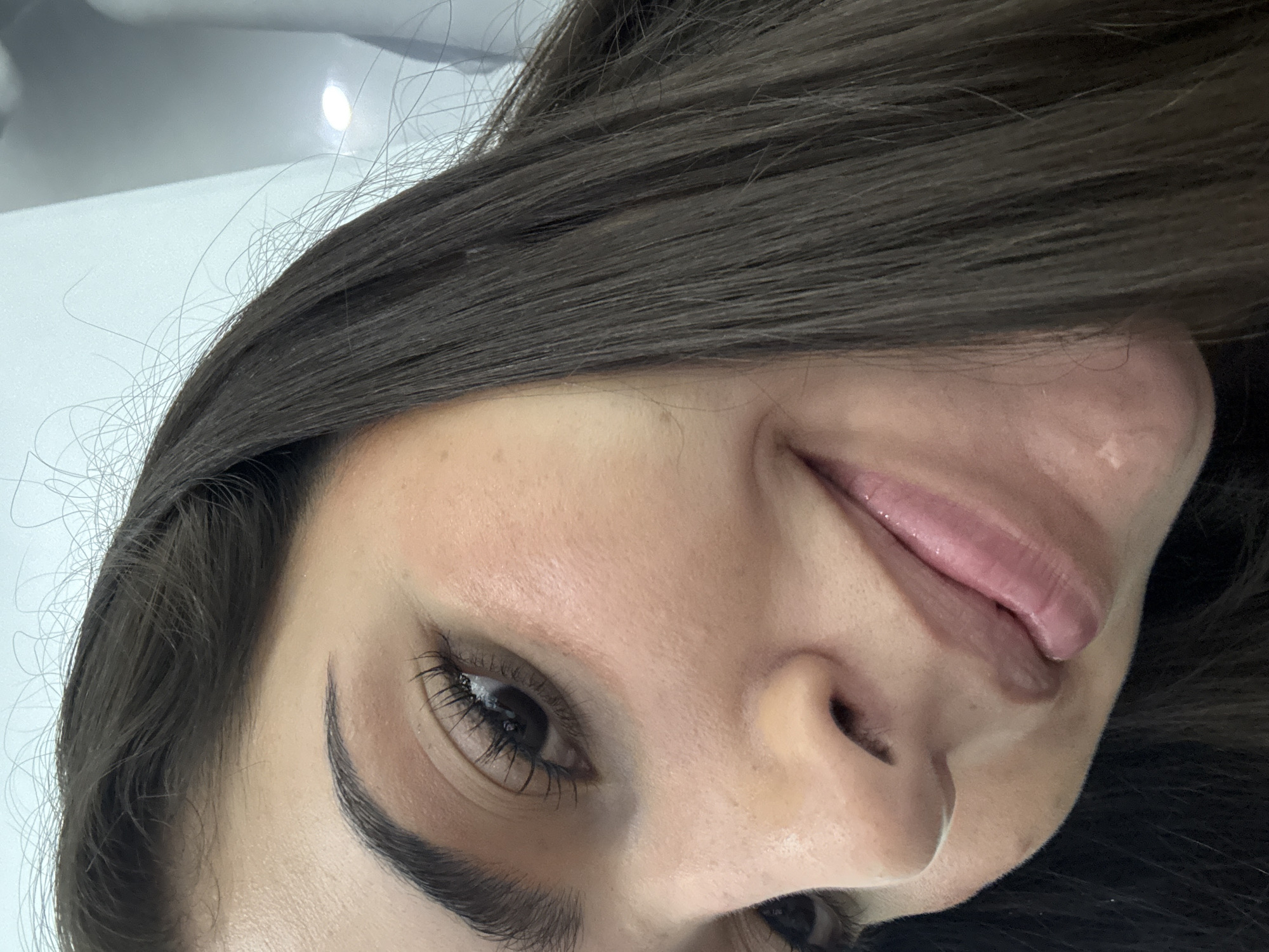 A close-up, rotated view of a woman's face. She has dark hair, dark eyes, and fair skin. Her left eyebrow is visible and well-defined. A small silver nose ring is present in her left nostril. Her lips appear to have lip gloss. The background is primarily a blurry white surface. It is impossible to determine if this image was taken in San Bernardino Tlaxcalancingo, Mexico without additional information.