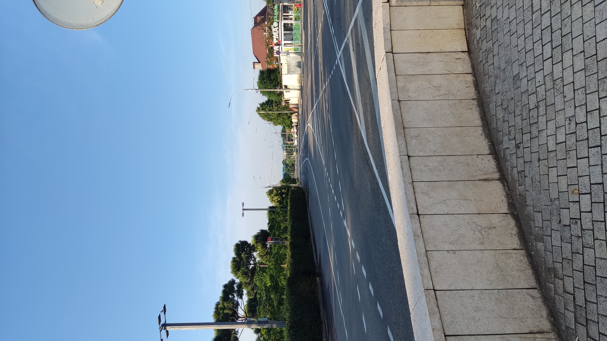 The image depicts a wide, multi-lane asphalt road in Krasnodar, Russia, under a clear blue sky. On the left side of the road, a well-maintained green hedge lines the perimeter, with tall pine trees and manicured bushes interspersed. A lamppost stands tall beside the hedge. The road itself is marked with white dashed and solid lines, indicating multiple lanes and directions of traffic. In the distance, several cars are visible, suggesting moderate traffic. On the right side of the road, a low-rise building with a red-tiled roof houses a business with signage that appears to be in Cyrillic, possibly a small shop or cafe. Streetlights are spaced along the road, and traffic signals are present. In the foreground, a low, light-colored stone wall separates the road from a pedestrian walkway paved with gray rectangular stones. The overall scene is one of an urban environment, likely a main thoroughfare, captured during daylight hours.