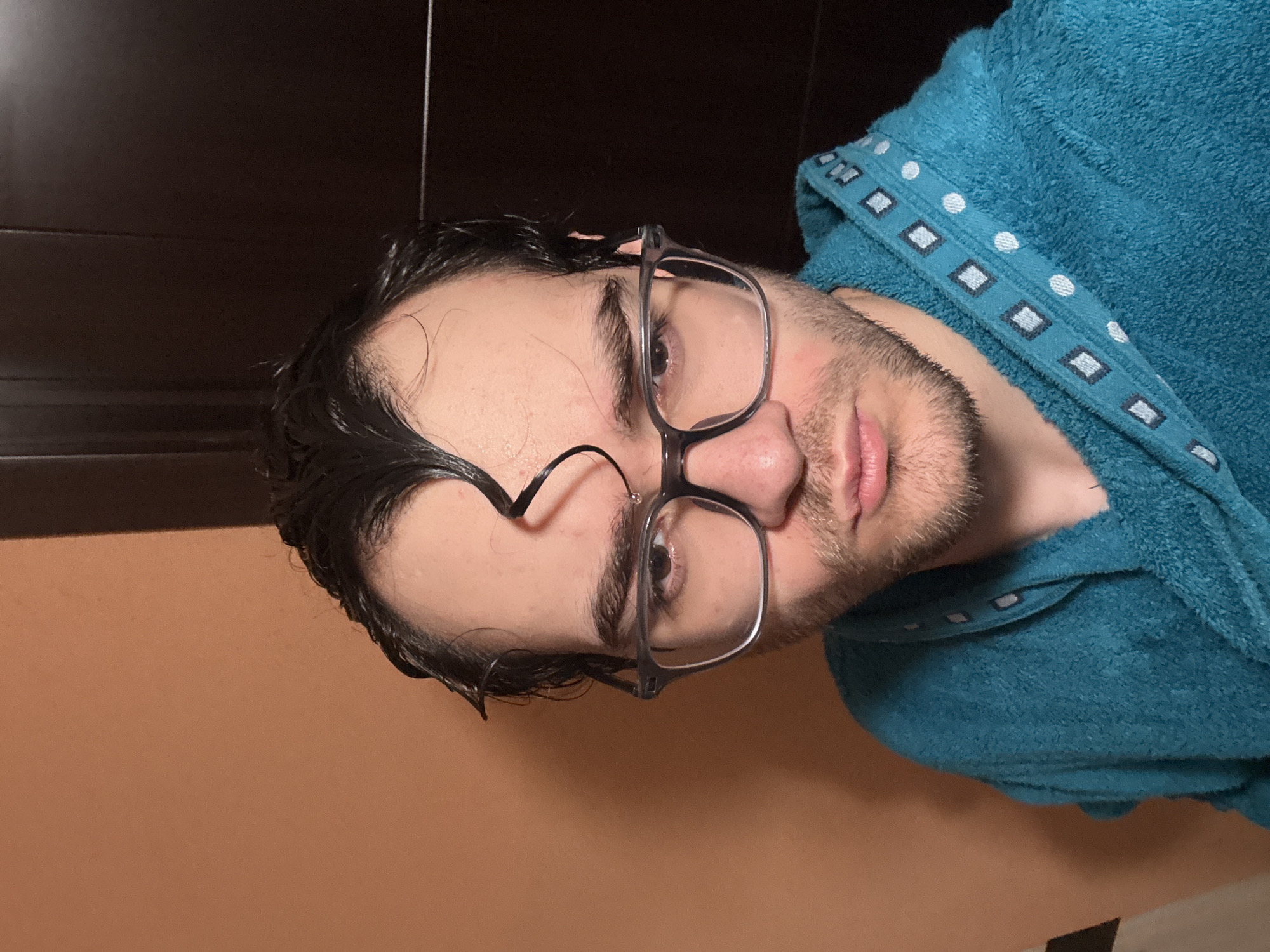 A selfie of a man with wet dark hair wearing glasses and a blue terrycloth robe. The background suggests an interior space, possibly a bathroom, with a two-toned wall – light orange on the bottom and dark brown at the top. The exact location in Blagoevgrad, Bulgaria, cannot be determined from the given image.