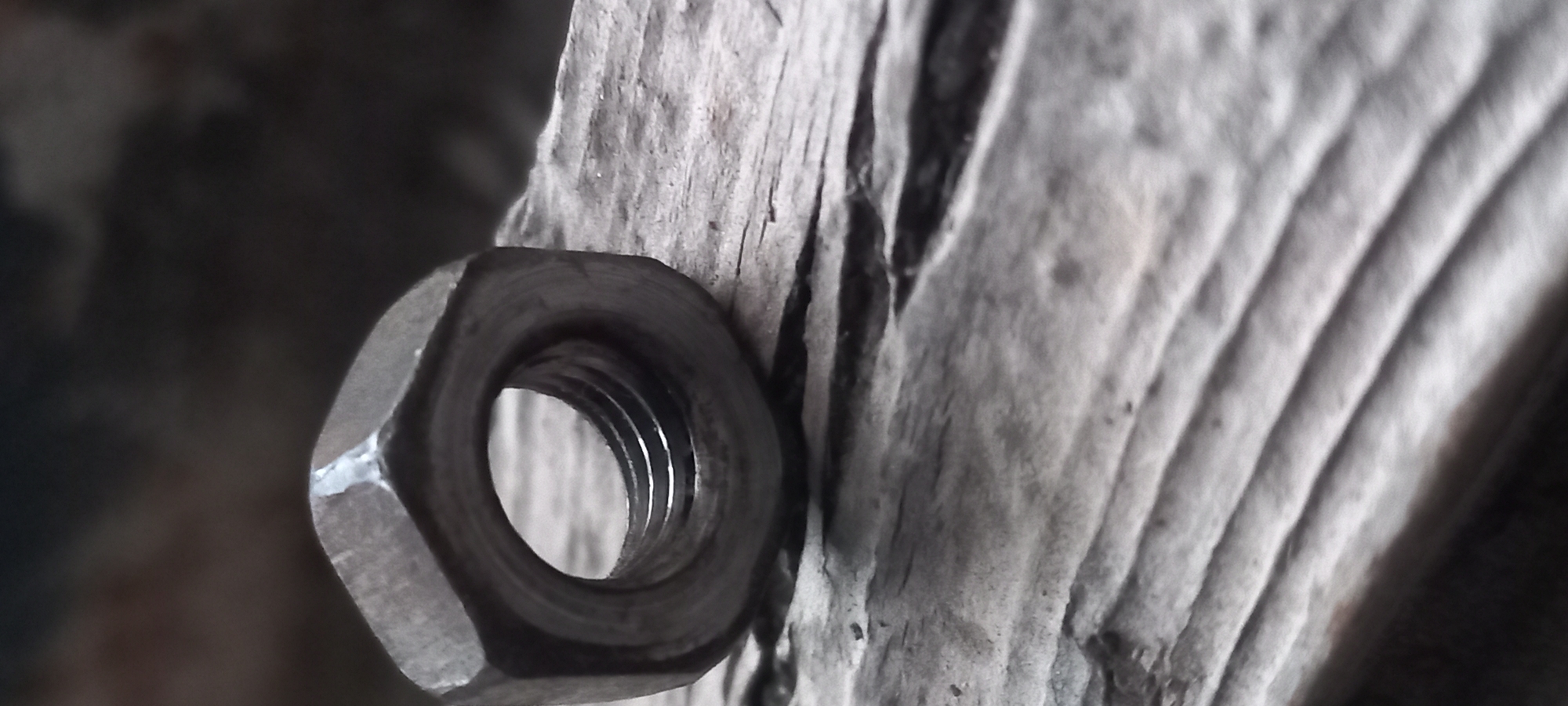 A weathered, hexagonal metal nut with a threaded interior rests on a textured, light-colored wooden surface in the city of Kryvyi Rih, Ukraine. The nut is positioned horizontally, revealing its dark grey metallic composition and the visible internal threading that catches reflections of light. The wooden surface displays prominent, roughly parallel grain lines and minor surface irregularities, indicating a natural and possibly aged material. The background is dark, out of focus, and indistinct, suggesting a shadowed environment. No individuals, actions, or specific interactions are depicted.