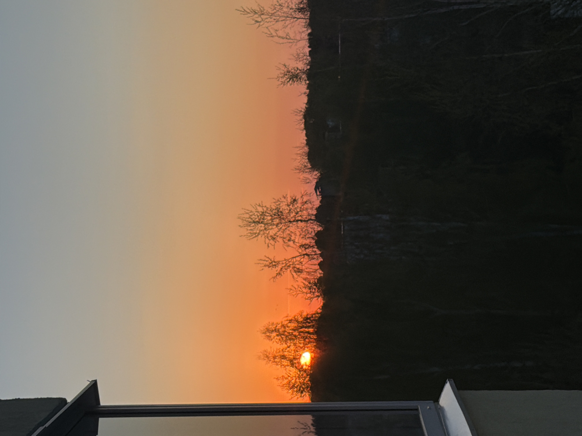 The image shows a sunset in what appears to be a rural area near Chaikovsky, Russia. The sky is gradient, transitioning from a pale gray-blue to a warm orange near the horizon. The sun is visible, partially obscured by trees, casting an orange glow. Silhouetted trees and some low structures are visible along the horizon line. In the foreground, there is a portion of a window and a building structure.