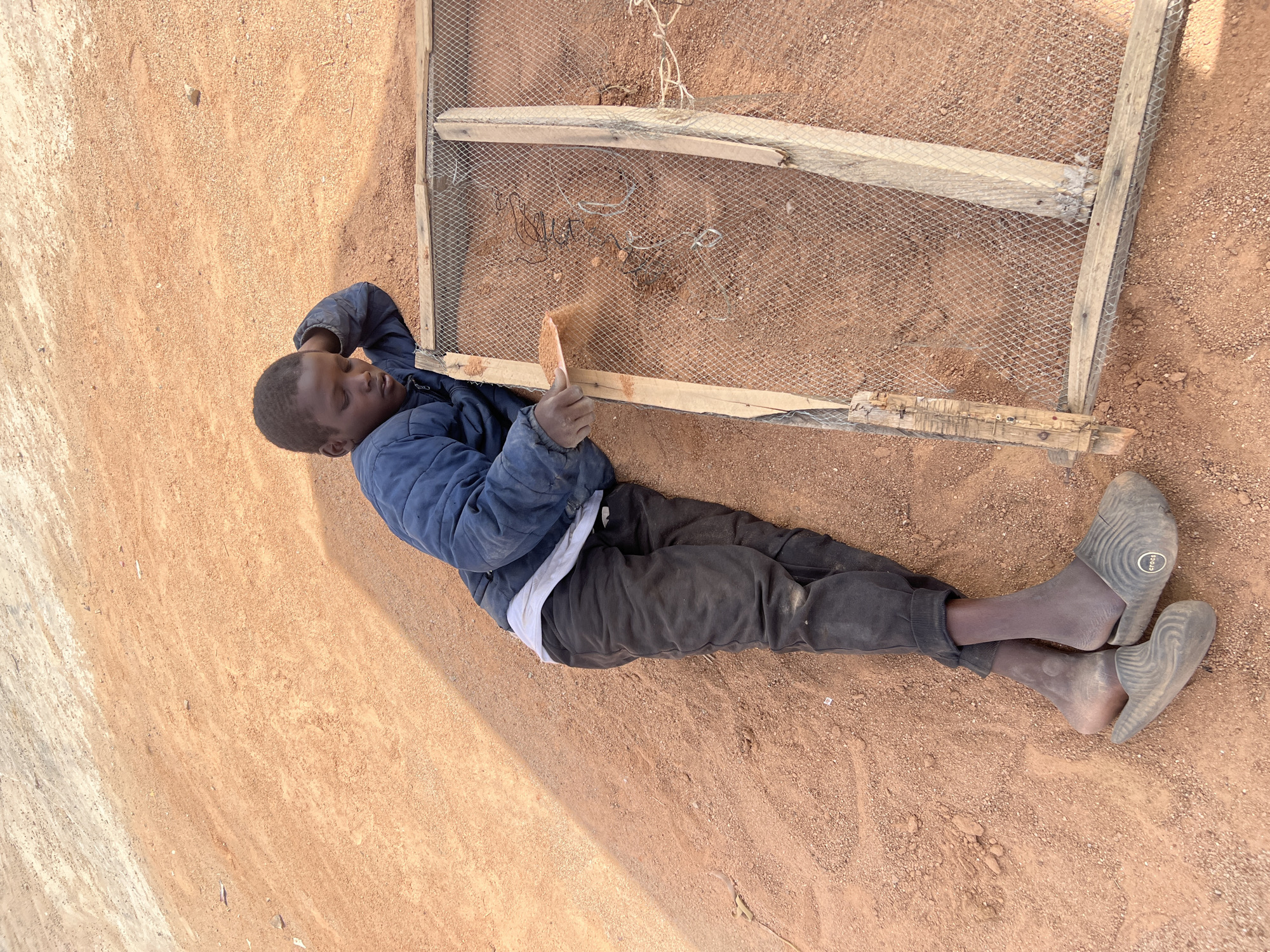 A youth lies on the ground in Barakin Lerawi, Nigeria, sifting dirt through a wire mesh frame using a small trowel. He wears a blue jacket, black trousers, a white shirt, and gray Crocs shoes. The ground is reddish-brown dirt.