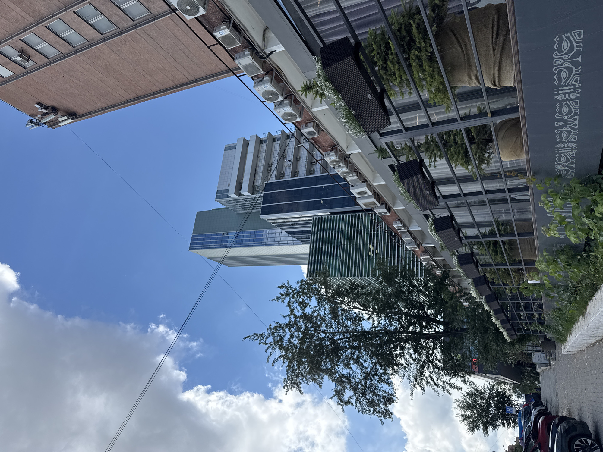 This media file depicts a street scene in Yekaterinburg, Russia, likely during the daytime. The perspective is upward-facing, looking towards several buildings and the sky.
In the foreground, an overhead structure with a dark, textured surface is visible, adorned with plants and white flowers. Below this structure, a row of parked cars is visible along the side of the street, suggesting an urban or commercial area.
Towering in the background are modern buildings, some with glass facades and others with a more traditional brick exterior. A large tree with dense foliage is also prominent, its branches reaching towards the sky. The sky is blue with scattered white clouds, indicating fair weather.
Several air conditioning units are mounted on the exterior of the buildings, a common sight in urban environments. There is no discernible activity or event taking place, suggesting a still or observational shot. No text is clearly visible within the frame, though a parking sign may be present near the cars. The overall impression is that of a city landscape on a clear day.