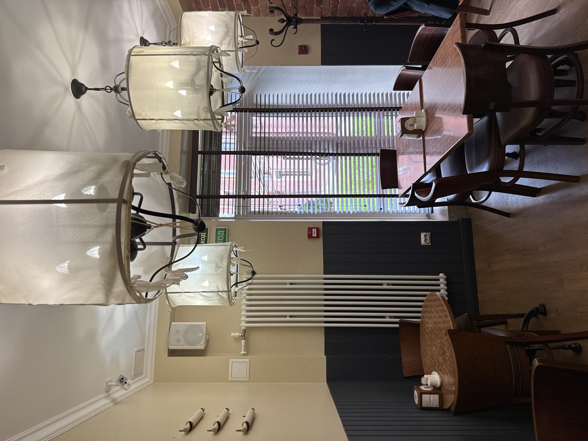 The image shows the interior of a restaurant in Saint Petersburg, Russia. The room is furnished with wooden tables and chairs, and illuminated by several large, fabric pendant lamps. Two tables are visible in the foreground, one round and one rectangular. The walls are painted a pale yellow, with a dark blue wainscoting. A large white radiator is mounted on the wall. To the right, a tall coat rack stands near a brick wall. Through a window with horizontal blinds, a glimpse of the exterior can be seen, with brick buildings and greenery. Two green exit signs with Russian text are visible hanging from the ceiling.