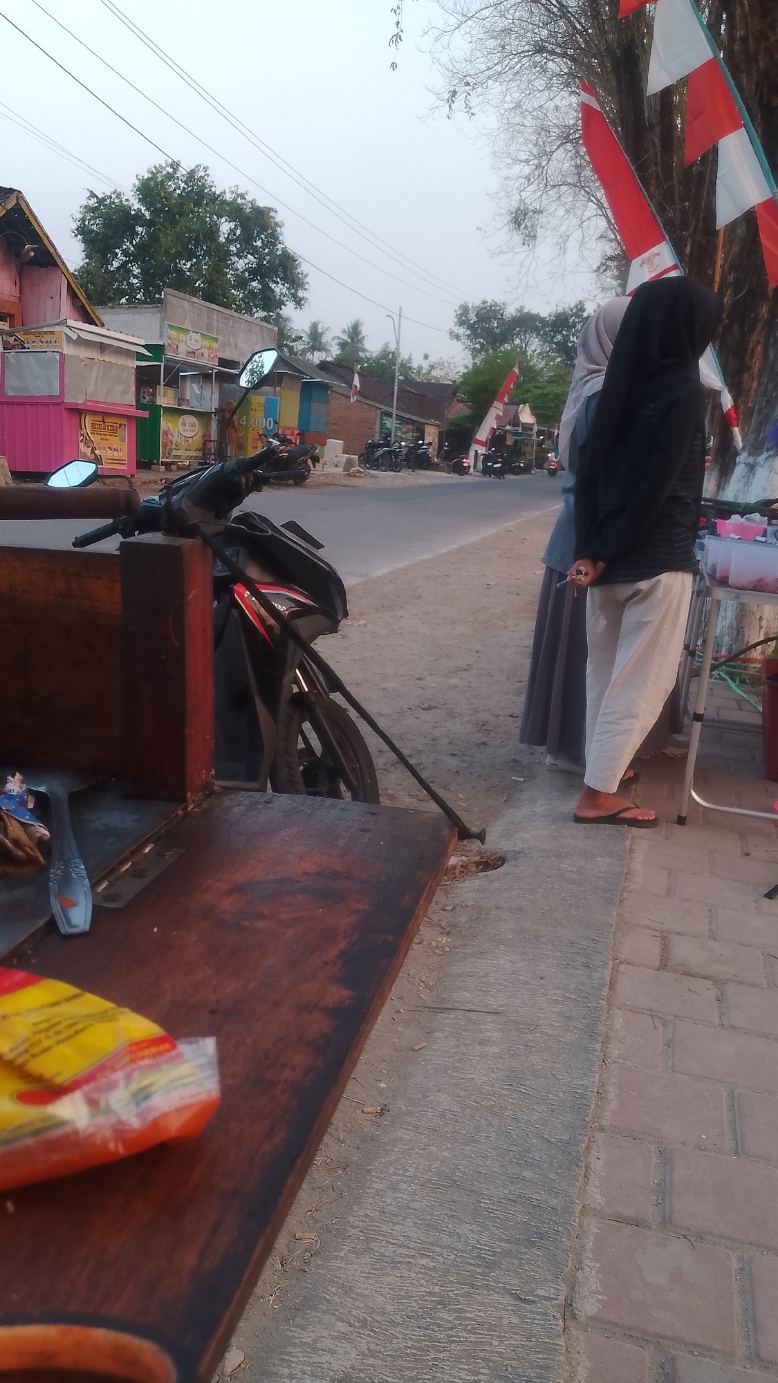 The image depicts a street scene in Karanglangu, Indonesia, with several stalls and parked motorcycles. In the foreground, a wooden cart or food stall is visible, with a silver spoon on its surface and a yellow and orange packaged item partially obscuring the wood. To the right of the cart, a black motorcycle is parked, leaning on its kickstand. Further down the street, two individuals, likely women, are standing. One is wearing a black hijab and a black long-sleeved shirt with white pants, and the other is wearing a light blue hijab and a grey long skirt. They appear to be interacting with a vendor at another stall, which features clear plastic containers with red contents. Several other motorcycles are parked along the roadside, and small Indonesian flags are attached to trees and poles. The environment suggests a rural or semi-urban setting with a paved road and a sidewalk made of bricks and concrete. The lighting indicates it is either early morning or late afternoon.