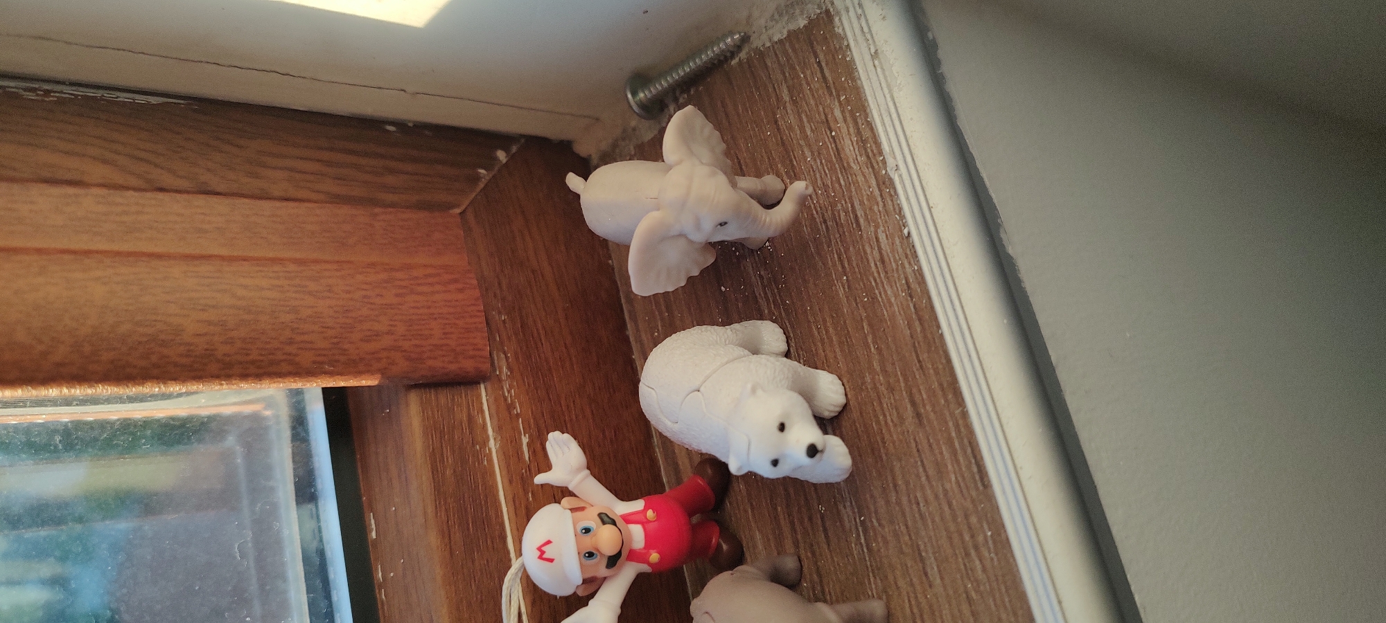 On a surface resembling wood grain, next to a window frame, are several small, toy-like figures. These include a white elephant, a white polar bear, and a Mario figurine wearing a white hat with a red "M" on it and a red outfit. A screw is visible near the elephant.
