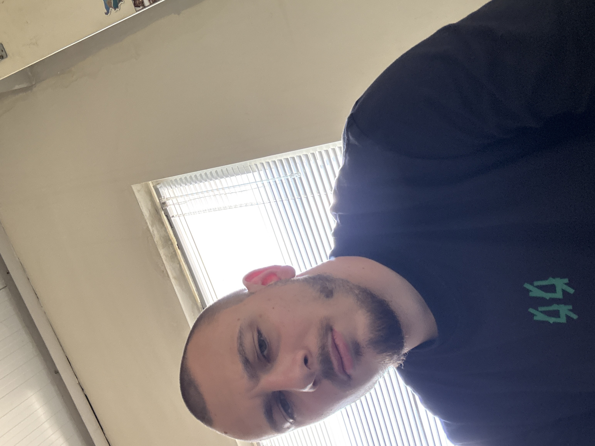A close-up selfie is taken from a low angle, focusing on the face of a young adult male. He has a shaved head, light stubble on his jawline, and a small goatee. His skin is fair, and he appears to be looking directly at the camera with a neutral expression. He is wearing a dark t-shirt with a green graphic design of what appear to be stylized numbers or symbols on the left chest area. The background consists of a beige wall and a window with horizontal blinds, allowing natural light to enter the room. Part of a white ceiling is visible at the top of the frame. The image lacks any specific environmental cues to pinpoint a location within Stara Zagora, Bulgaria, other than the fact that it is an indoor setting.