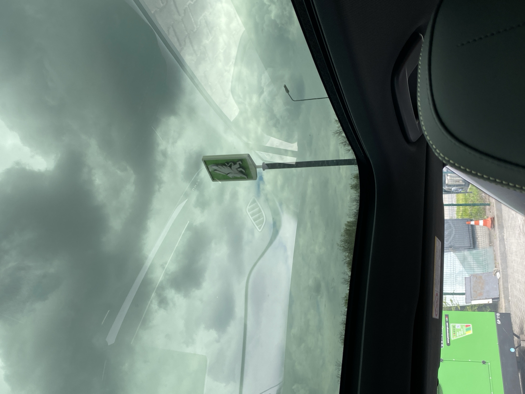 The image shows a view from inside a vehicle, looking out through the windshield. The view is rotated clockwise. Outside, a road sign bearing an image of a white unicorn on a green background is visible. A street lamp is also visible in the background. In the foreground, parts of the car's interior, including the windshield, dashboard, and the side of a car seat, are visible. On the right side of the frame, an electric generator with a bright green panel is visible. The location is an industrial area with fencing, a safety cone, and other industrial objects. The city is unknown.