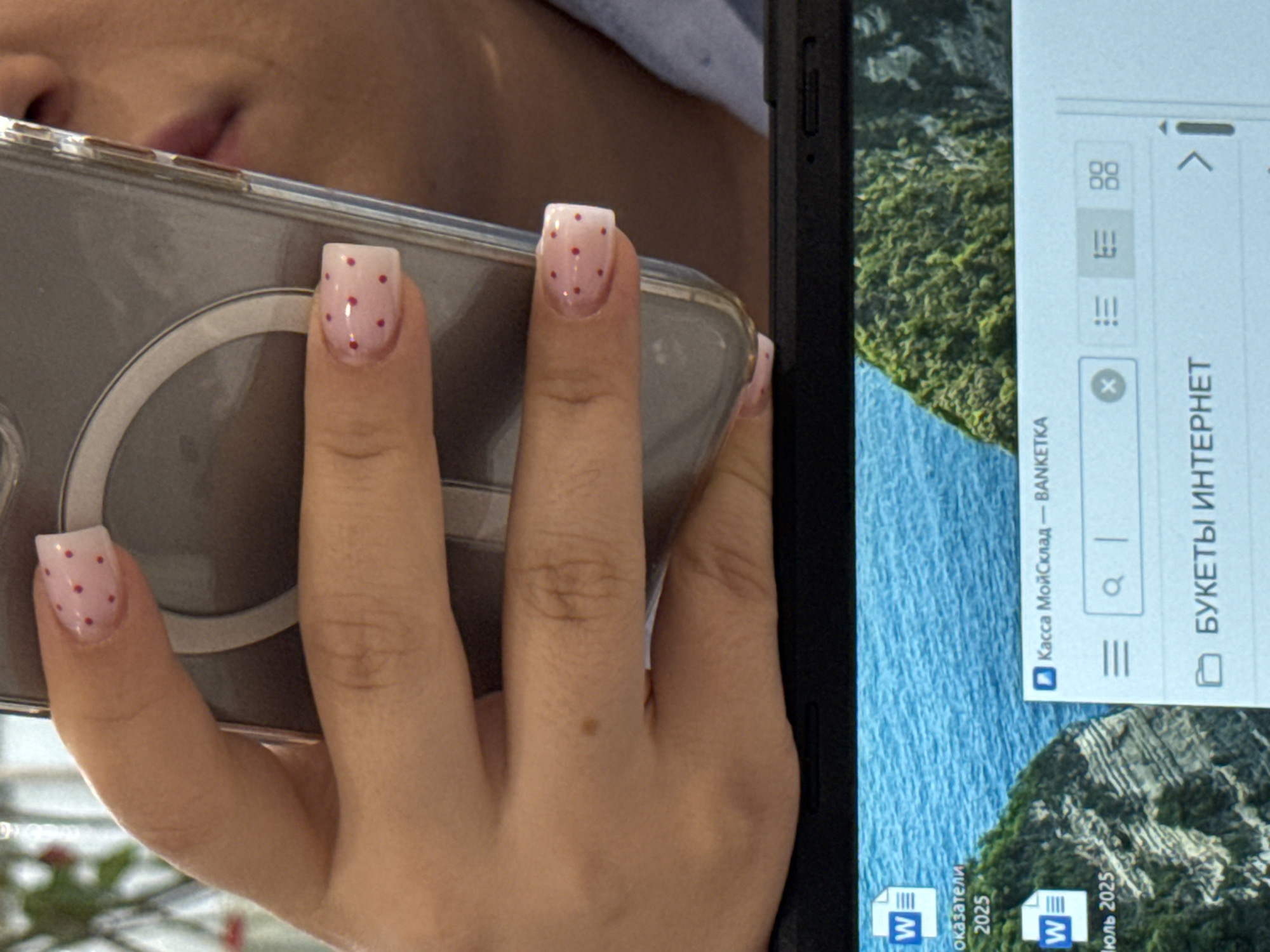 A close-up, slightly angled shot shows a person's left hand with light pink, square-tipped nails adorned with small red dots. The hand is holding a transparent phone case, revealing the back of a phone with a circular cutout. In the background, a portion of a person's face is visible. Below the hand and phone, a laptop screen displays a desktop with several icons, including two "W" icons for Microsoft Word documents labeled "оказатели 2025" and "июль 2025." A file explorer window is open, showing a title "Касса МойСклад — BANKETKA" and a folder icon labeled "БУКЕТЫ ИНТЕРНЕТ". The background of the desktop is a scenic image of a rocky coastline with lush green vegetation. The image is set in the city of Assinovskaya, Russia.