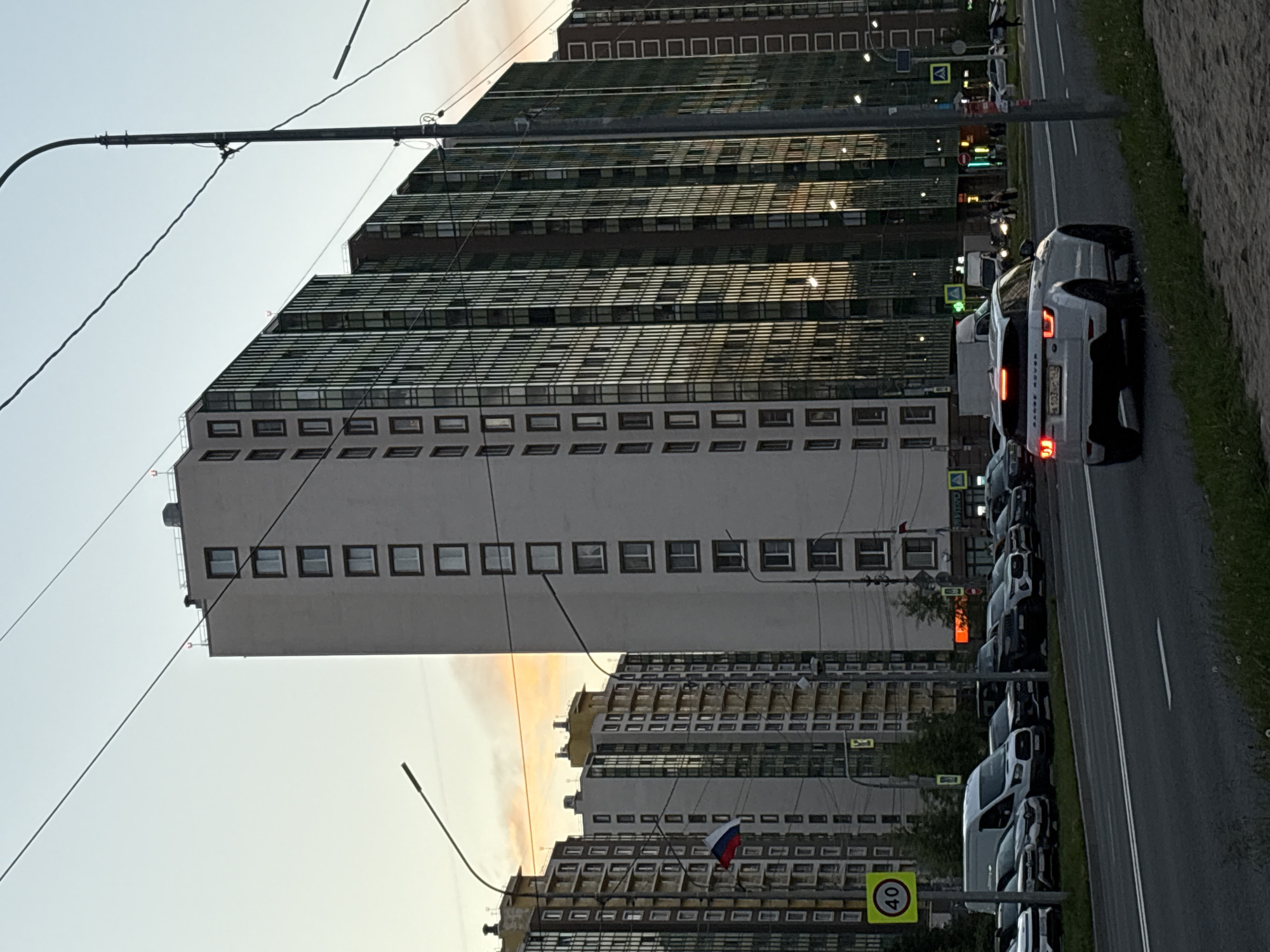 A white Range Rover is seen driving on a city street in Russia. The car is in the foreground, with its taillights illuminated. In the background, several tall apartment buildings are visible. One building is white with many windows, and another is covered in reflective glass. A Russian flag is hanging from a pole near one of the buildings. The sky is a pale orange and blue, suggesting it is either sunrise or sunset. A speed limit sign indicating 40 is visible on the left side of the image, along with a row of parked cars and vans. Overhead wires and a streetlamp are also present in the scene.