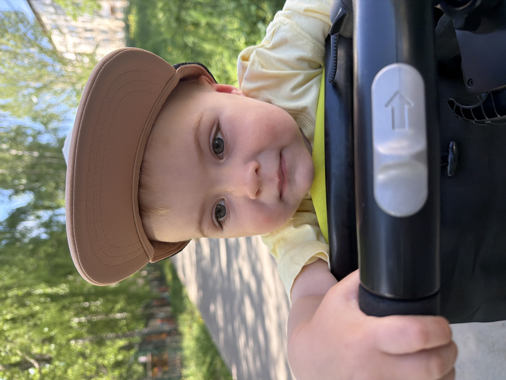 A young child is photographed outdoors on a sunny day. The child is wearing a tan cap and a light yellow long-sleeved shirt, and is positioned in a stroller, holding onto the black stroller handle. The background is slightly blurred and shows greenery, including trees and a paved path. Buildings are also visible in the background. Specific location in Votkinsk, Russia, cannot be determined based solely on the visual information provided.