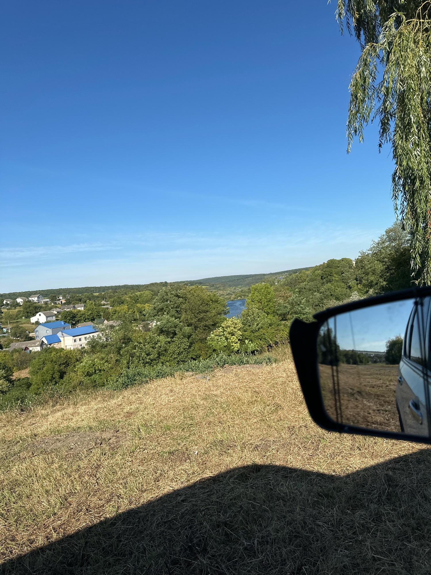 The image captures a view from a vehicle, looking towards a rural landscape in the city of Makiv, Ukraine. In the foreground, dry, sun-bleached grass covers a sloping hill, casting a distinct shadow from the vehicle. To the right, a side-view mirror reflects a portion of the scene, showing a field and trees. In the middle ground, a cluster of houses with blue roofs are visible amidst lush green trees. Beyond the houses, a body of water, likely a river, winds through a valley bordered by rolling, tree-covered hills under a clear, bright blue sky. A weeping willow tree extends into the upper right corner of the frame, its long, green branches hanging down. The overall impression is one of a sunny, rural day.