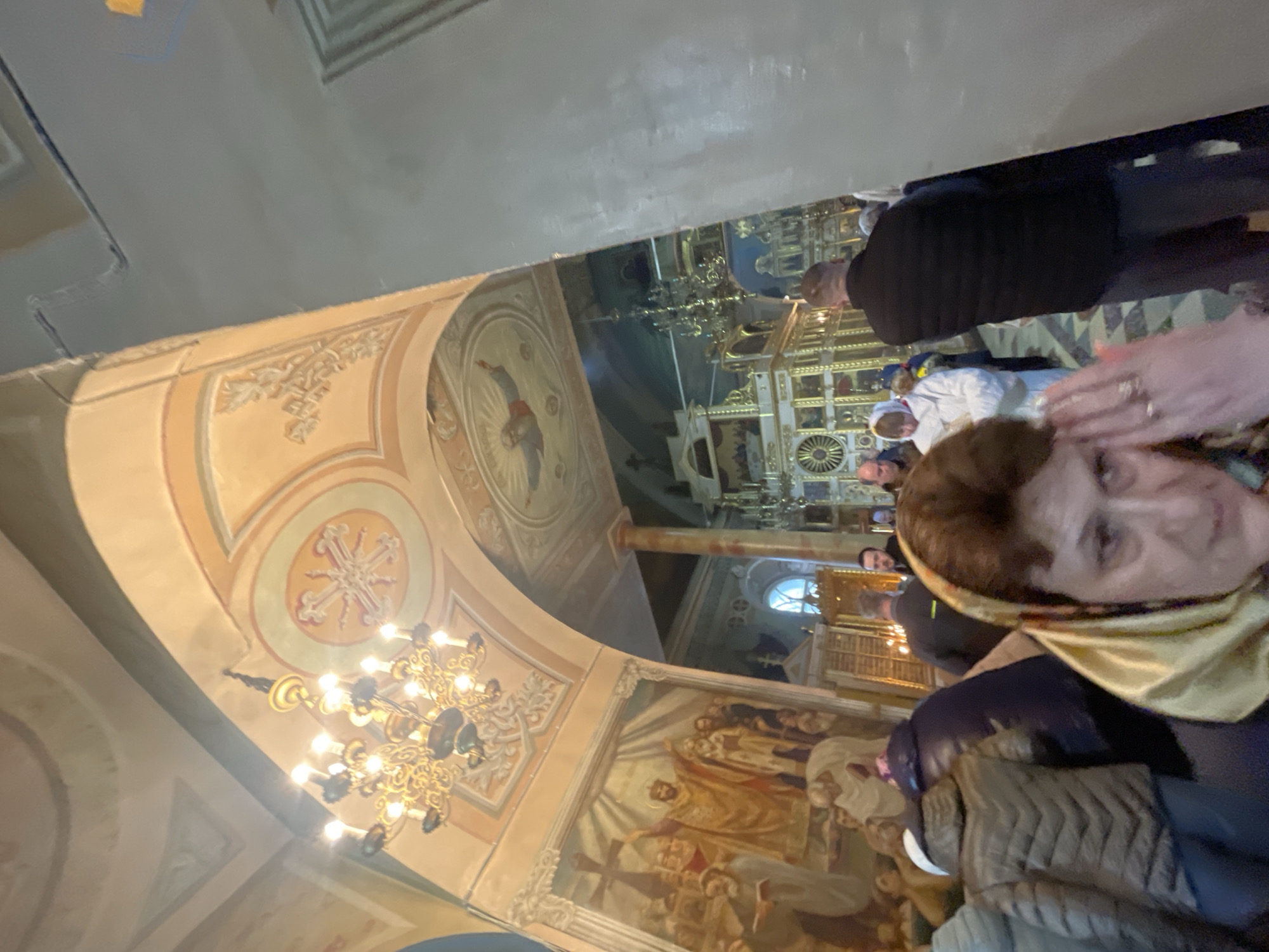 The image shows the interior of a church in Ivanisovo, Russia. It is shot from a low angle, showcasing the ornate ceiling, murals, and chandeliers. The walls are painted with religious scenes, and the ceiling features decorative designs. People are present, some appearing to be in prayer or contemplation. A woman with a headscarf is partially in the foreground, holding a camera.