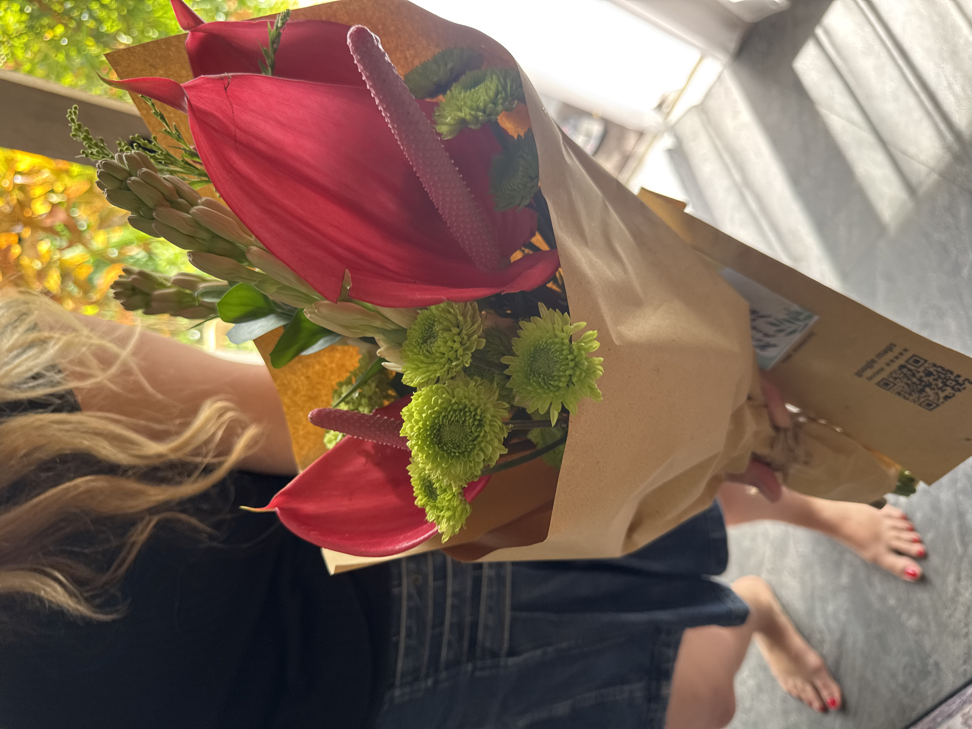 A woman is holding a bouquet of flowers in a paper bag. The bouquet contains red Anthurium flowers, green chrysanthemums, and what appear to be tuberoses. The woman is wearing a dark-colored shirt and denim shorts. She is kneeling on a gray floor that seems to be either stone or tile. The woman's blonde hair is partially visible. Her feet are bare with red nail polish on her toes. The paper bag has printed text on it, including the words "google maps" and a QR code. The location is likely in North Kuta, Indonesia. The image is lit by natural sunlight coming through a window. The foreground shows a blurred view of green foliage and yellow sunlight.