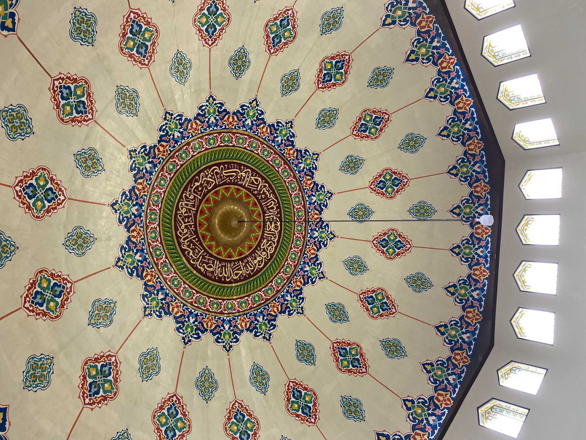 The image shows the ornately decorated interior dome of a mosque, likely in Lhokseumawe, Indonesia. The dome is painted with intricate geometric and floral patterns in vibrant colors including red, blue, green, and gold. A central medallion features Arabic calligraphy and a golden chandelier hangs from its center. Smaller decorative motifs are spread across the dome, radiating outwards from the center. Below the dome, a row of arched windows with decorative stained glass lets in natural light. The walls of the mosque are a neutral color. The overall impression is one of detailed artistry and spiritual ambiance.
