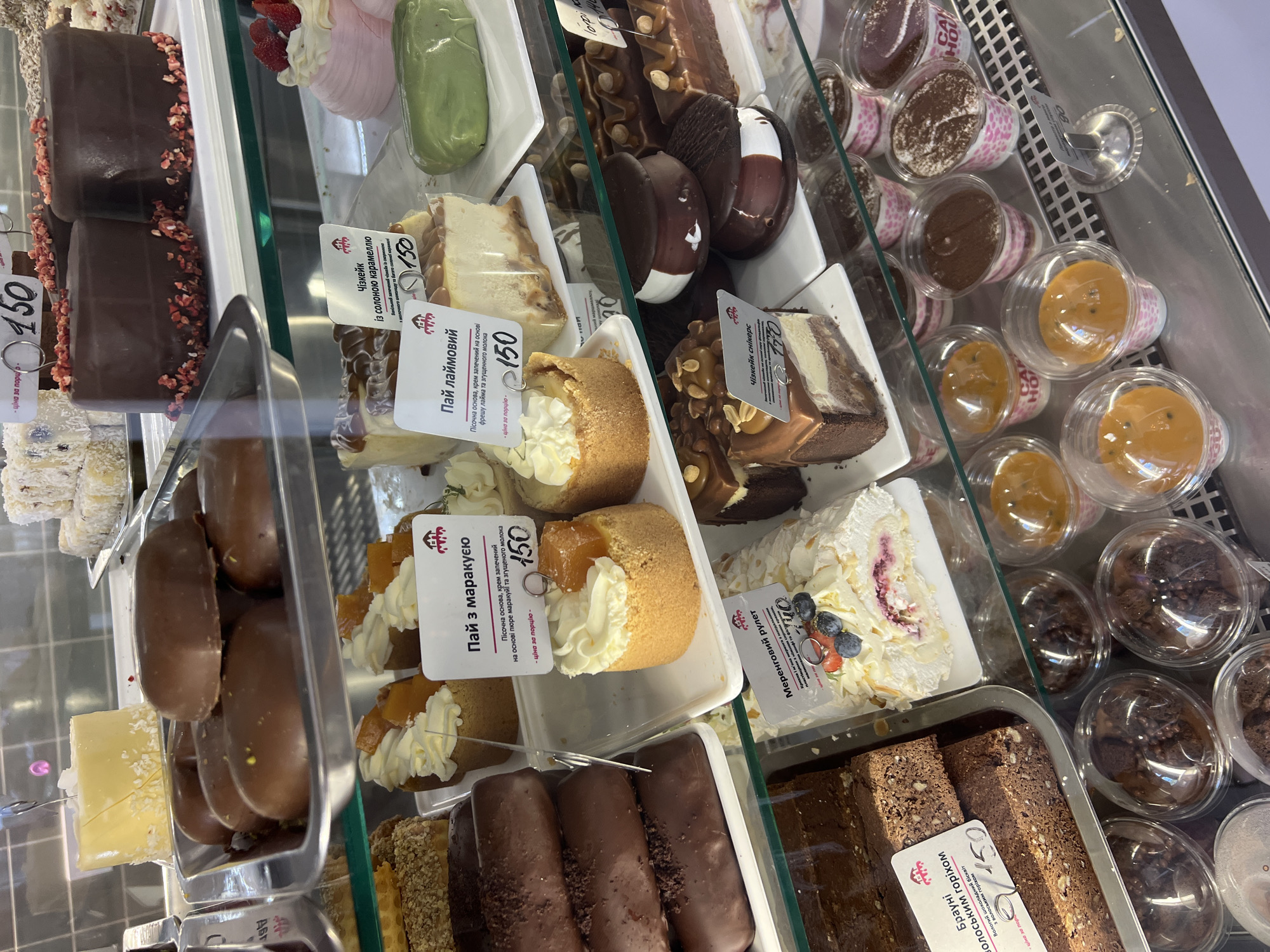 The image shows a refrigerated display case filled with various desserts, likely in a bakery or cafe in Odesa, Ukraine. In the top shelf, several chocolate-covered desserts are visible, some topped with sprinkles or fruit. Below them, a bright yellow cake is on display. The middle shelves feature individual cheesecakes with caramel and fruit toppings, small tart-like pastries with cream and fruit, and a green, oblong dessert. There are also chocolate cakes with nuts and chocolate icing, and what appear to be chocolate sandwich cookies. The bottom shelf holds chocolate brownies with nuts and individual cups of layered desserts, possibly mousse or tiramisu, and fruit-flavored desserts. The display case is made of glass and metal, and the overall environment appears clean and well-lit. Several price tags with Ukrainian text and prices in Ukrainian Hryvnia (UAH) are visible, indicating the location is a commercial establishment selling food.