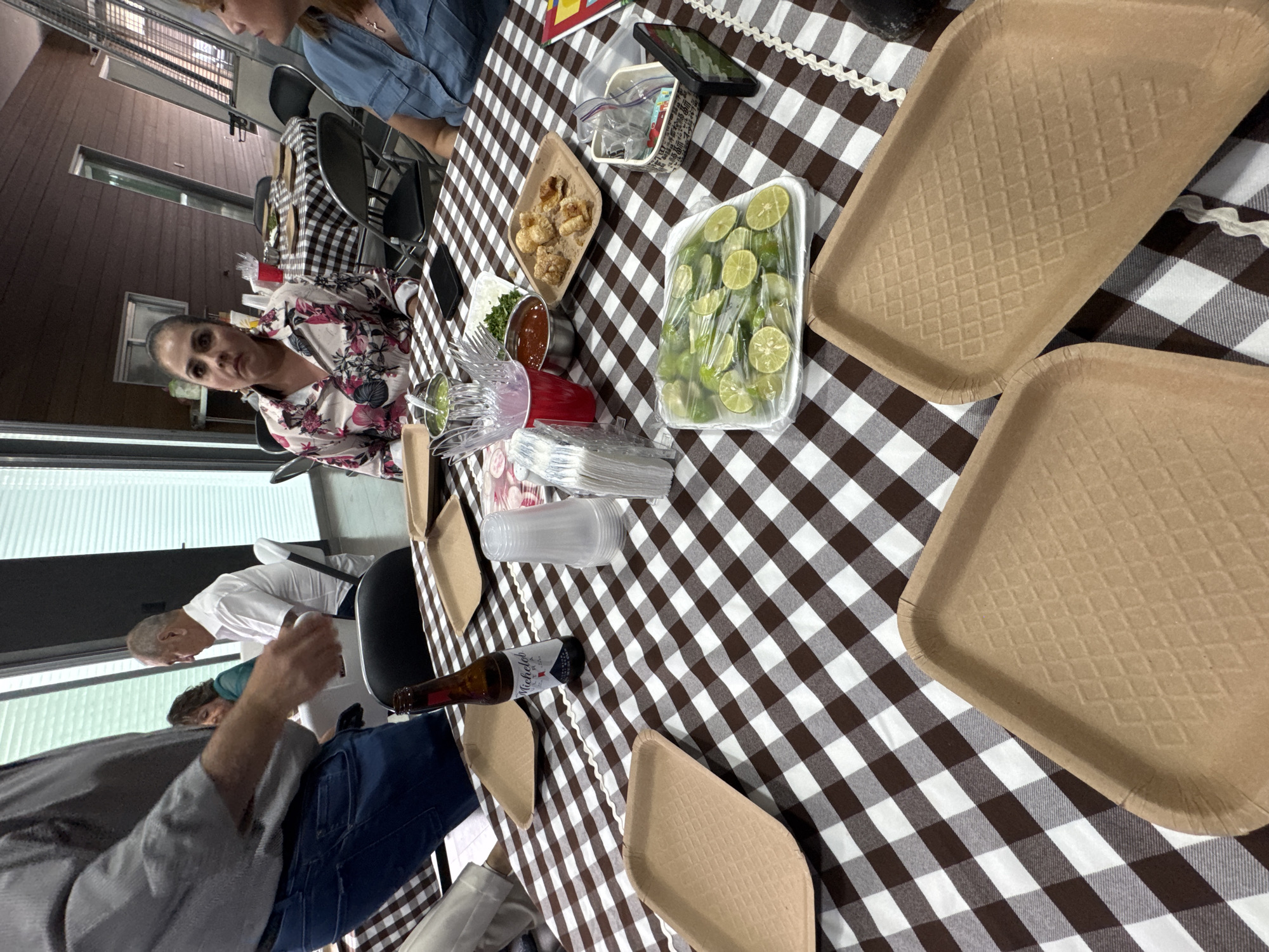 The image shows an outdoor gathering in San Bernardino Tlaxcalancingo, Mexico. A table covered with a brown and white checkered tablecloth holds a variety of items: food in disposable containers, including limes, fried pork rinds, and salsa; disposable cutlery and cups; several rectangular cardboard trays; and a bottle of beer. Three people are visible around the table. One woman with a floral top looks directly at the camera. Another woman with a denim top is partly visible. A man with a white shirt and an older man are also seated at the table. The background shows a building with windows and what appears to be a deck.