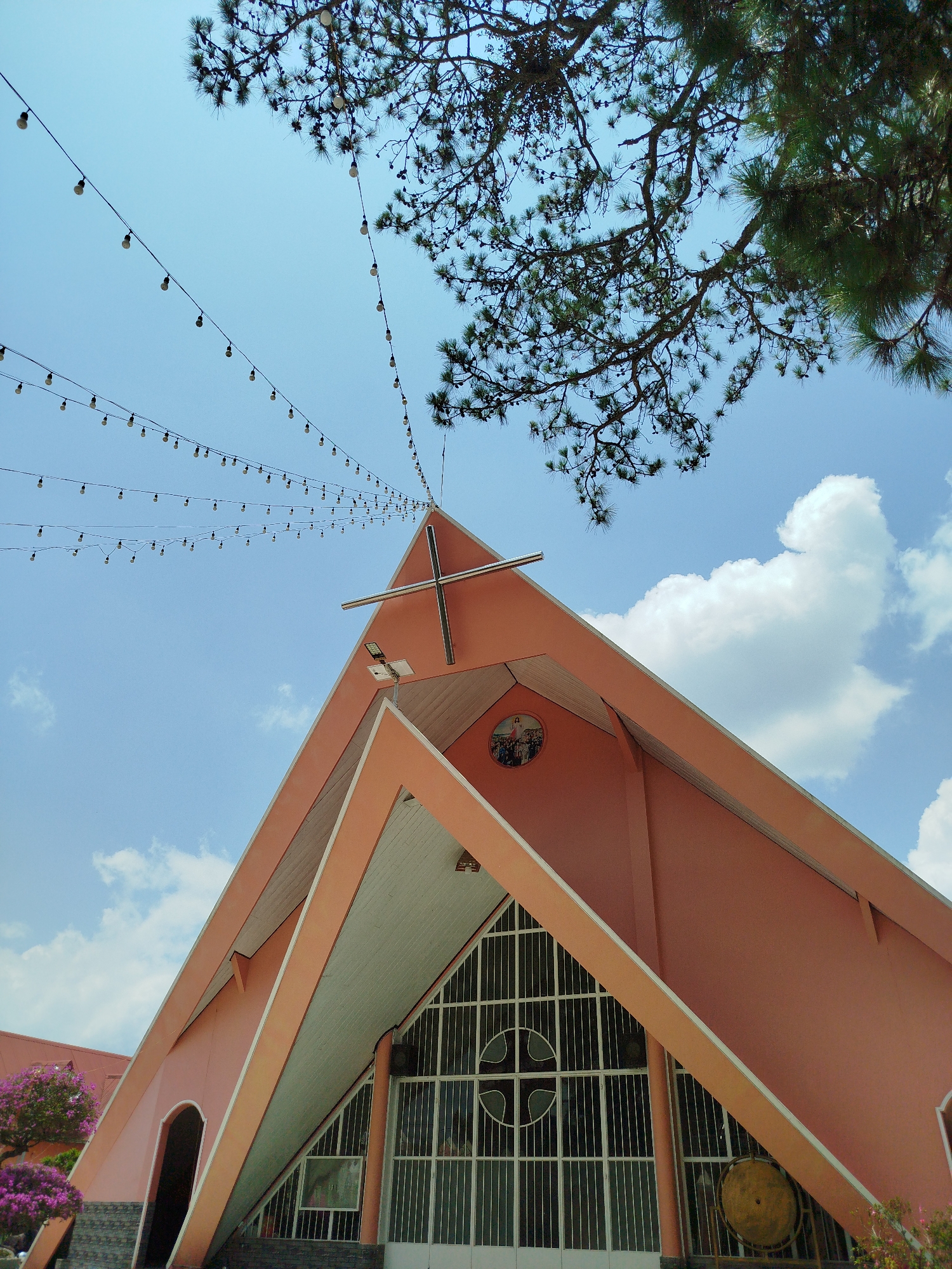 The image shows the facade of the Chicken Church (Nhà thờ Con Gà) in Đa Thọ, Vietnam, viewed from a low angle. The church is a salmon-colored building with a distinctive A-frame design. A large, silver cross is mounted on the peak of the roof. A string of light bulbs hangs from the top of the frame to the apex of the building. Part of a coniferous tree branch with pine cones is visible in the upper right. The sky is light blue with scattered cumulus clouds. The church has a large window with a cross design.