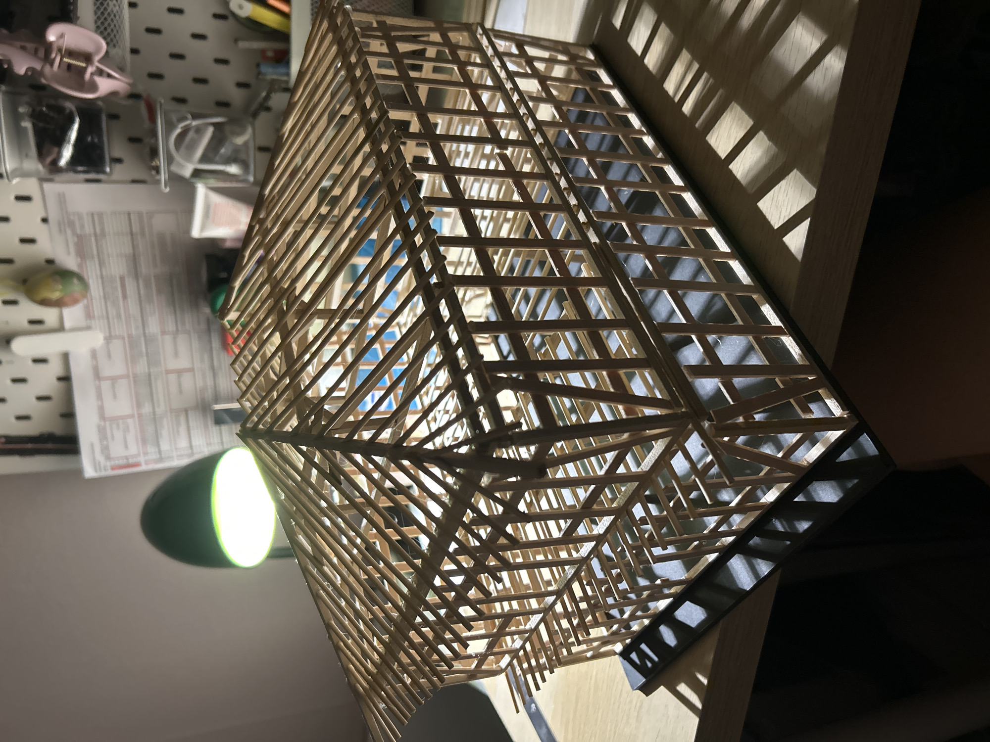 A detailed, forensic, matter-of-fact description of the scene is not possible. The image depicts a model of a wooden structure, possibly a house or a building, made of thin wooden sticks and beams. The model is placed on a wooden surface, and is illuminated by a green desk lamp, casting shadows on the table and the background. The background shows a pegboard with various items attached, including papers, containers with small objects, and a pink hair clip. There are no people visible in the image, and no specific location in the city of Sofia, Bulgaria, can be identified.