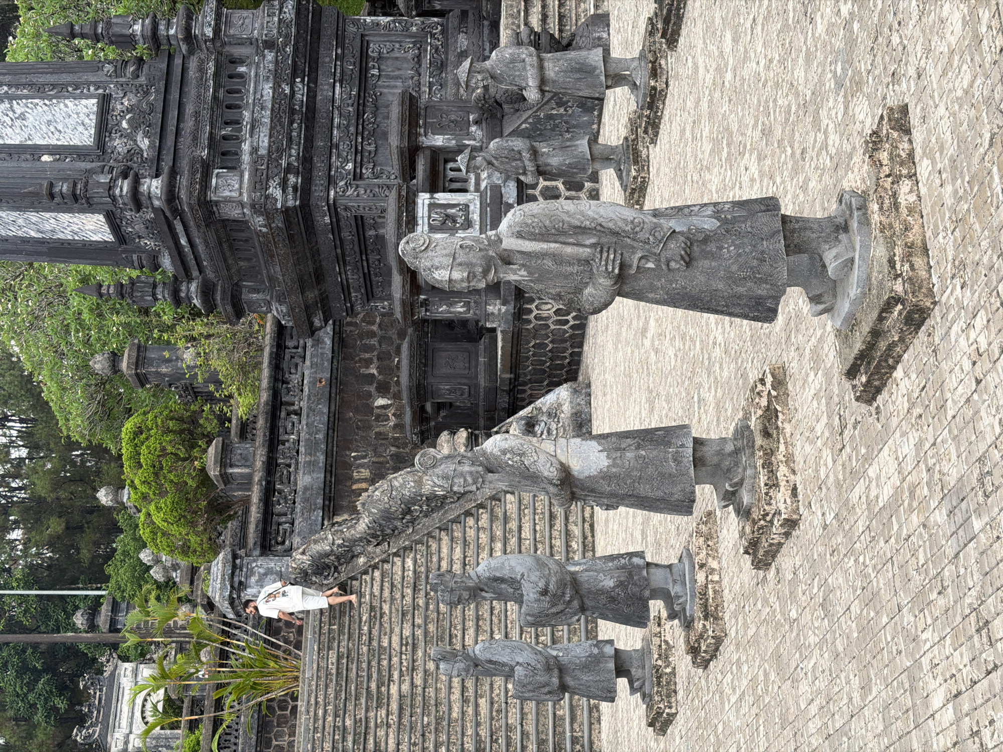 The image shows a vertical view of a stone stairway flanked by stone statues of figures in traditional dress. The statues line the stairway leading up to a stone building with intricate carvings and ornamentation. A man wearing white shorts and a white t-shirt is walking barefoot down the stairs. The scene is set amidst lush greenery.
The location is Thủy Xuân Ward, Vietnam.