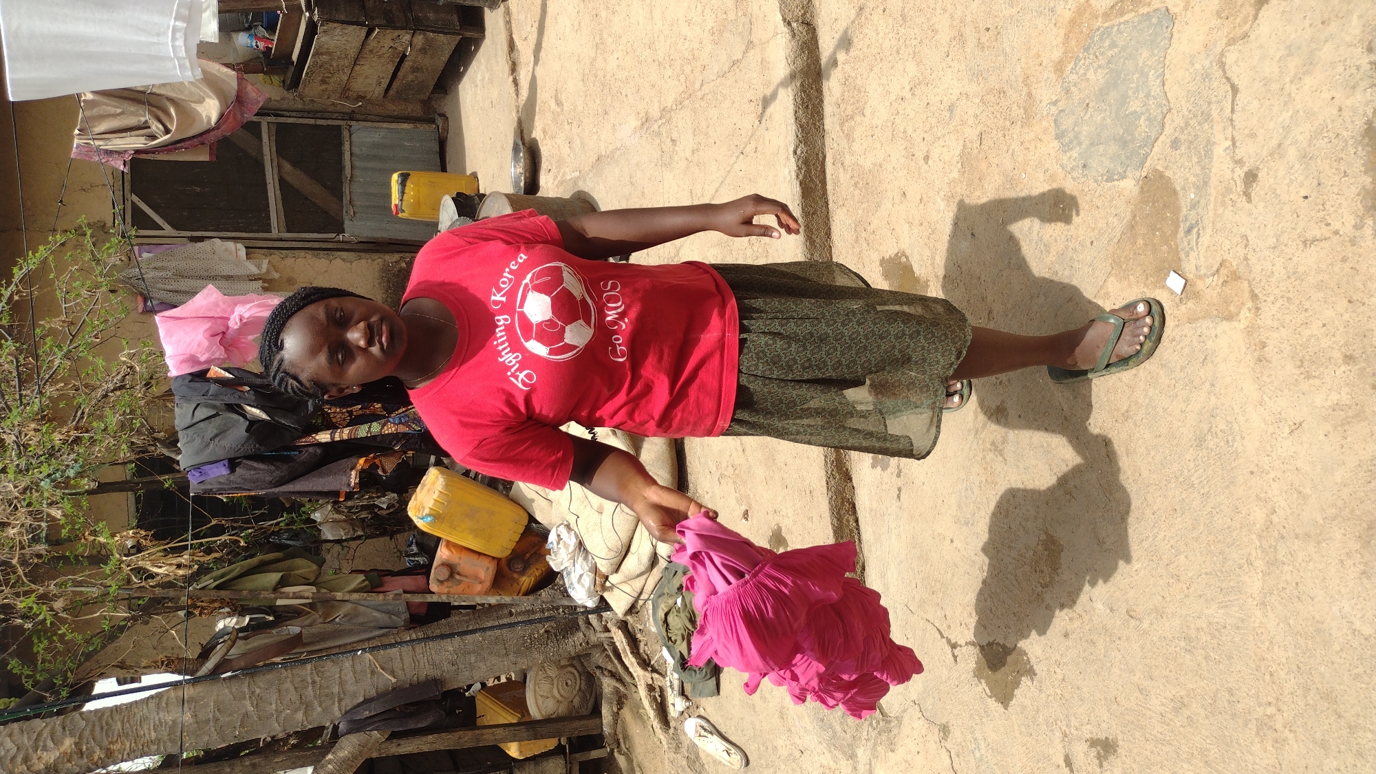 A woman stands in Jos, Nigeria on a cracked concrete surface outside a building, holding a pink garment. She wears a red t-shirt with a soccer ball design, a green patterned skirt, and green flip-flops. Her shadow stretches on the concrete. The area behind her is cluttered with various objects including yellow containers, drying clothes, and miscellaneous items.