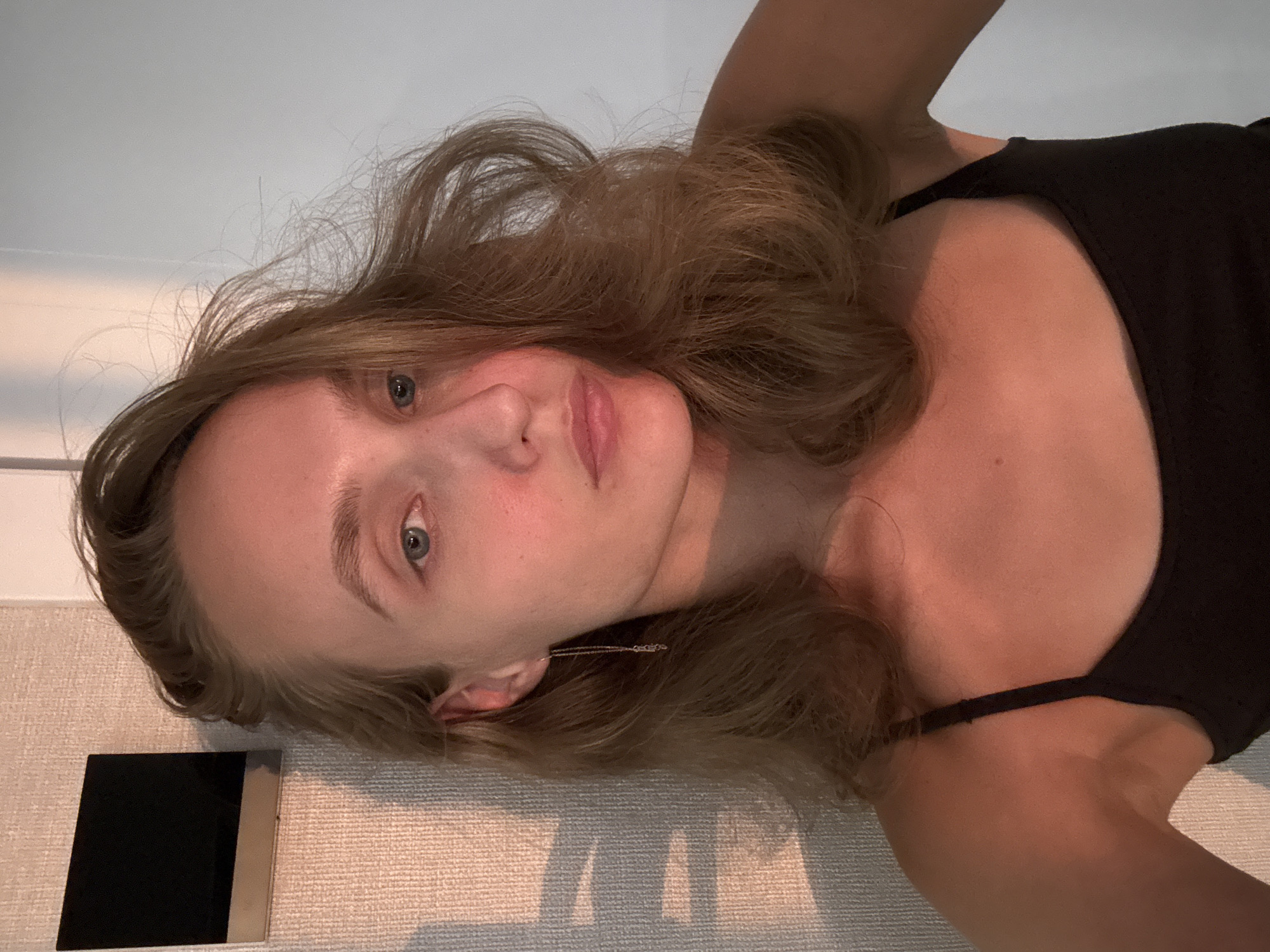 A young woman with blonde hair and blue eyes is captured in a close-up, selfie-style photograph. She is wearing a black tank top and a long, delicate chain earring on her left ear. The background consists of a textured wall with a dark square object mounted on it, and a plain white wall. Sunlight casts a warm glow on her face and skin, highlighting her features and creating shadows on the wall to her left. The overall impression is casual and intimate, possibly taken in a private living space or hotel room. There is no indication of the location being in the city of Podshivalovo, Russia.