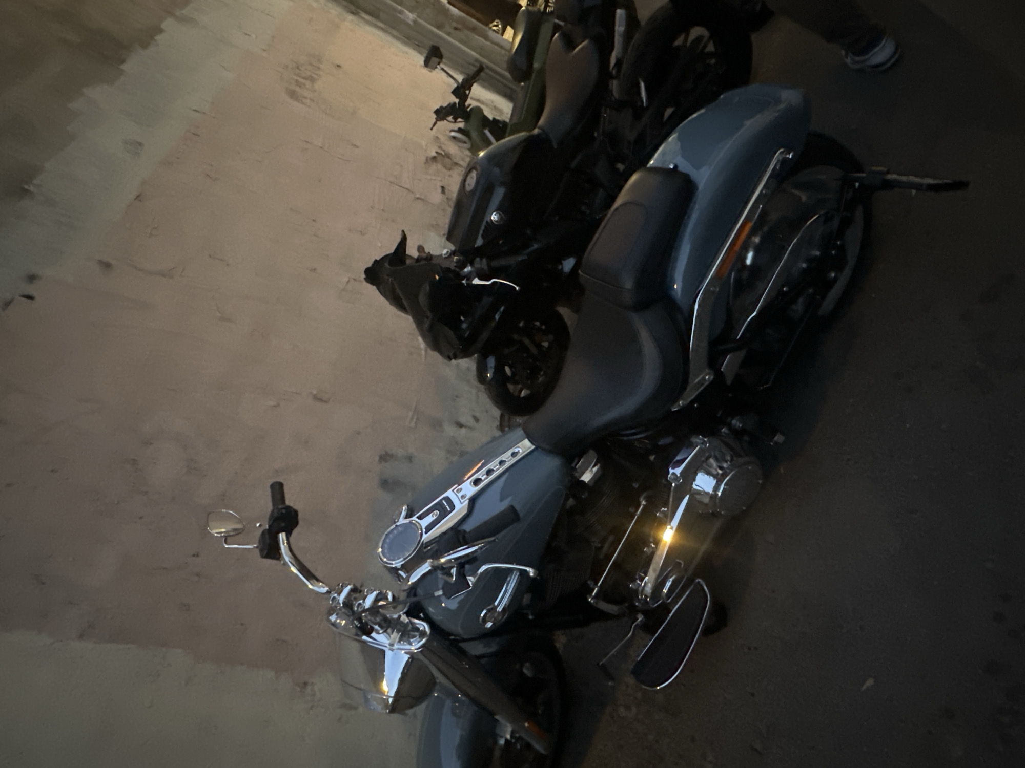 Two motorcycles are parked in front of a textured wall. The motorcycle in the foreground is gray and has chrome accents. The motorcycle in the background is black. A person's legs and feet are visible on the right side of the image, wearing athletic shoes. The scene appears to be outdoors in an urban environment, possibly a street or alleyway, with the lighting suggesting it is either dusk or night. There is no visible indication of the location being in Moscow, Russia.