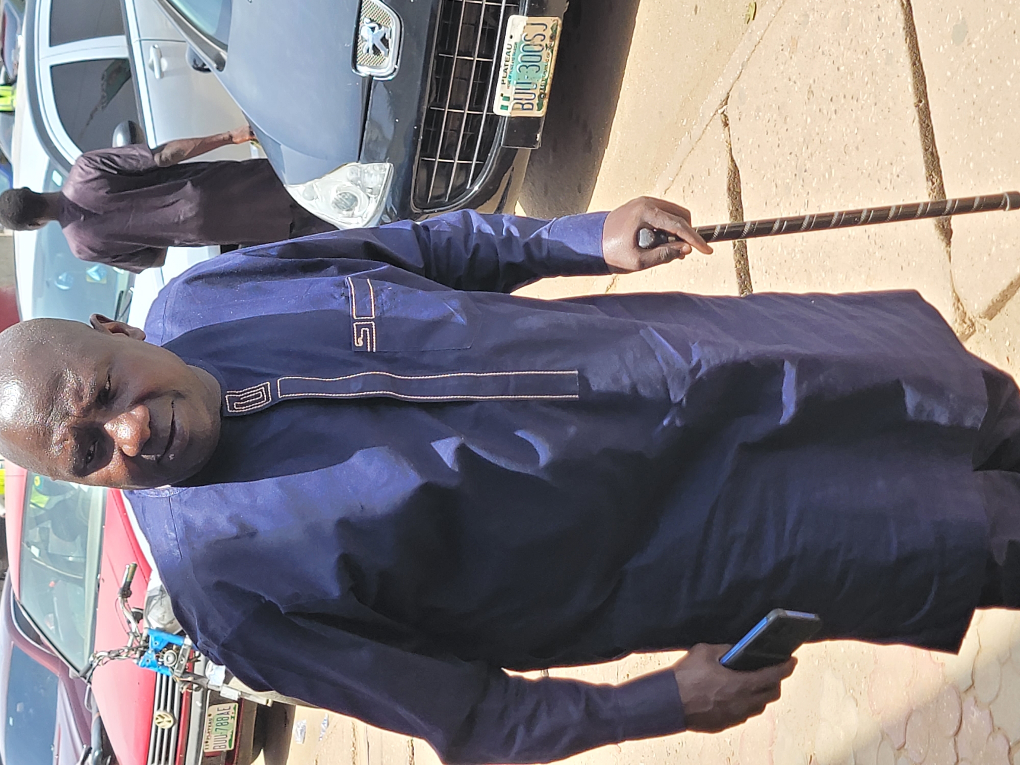 The image shows a man in a blue garment holding a cell phone and a cane while standing in what appears to be a street. A man wearing a brown shirt is standing next to a silver car. The license plate of a black Peugeot vehicle reads "BUI 300SJ" and indicates the car is registered in the Plateau State. Another vehicle, a red Volkswagen is parked nearby with the number plate "BUI 788AE" also indicating registration in Plateau state. Given that Plateau state is located within Nigeria, it is reasonable to assume this image was taken in the city of Jos.