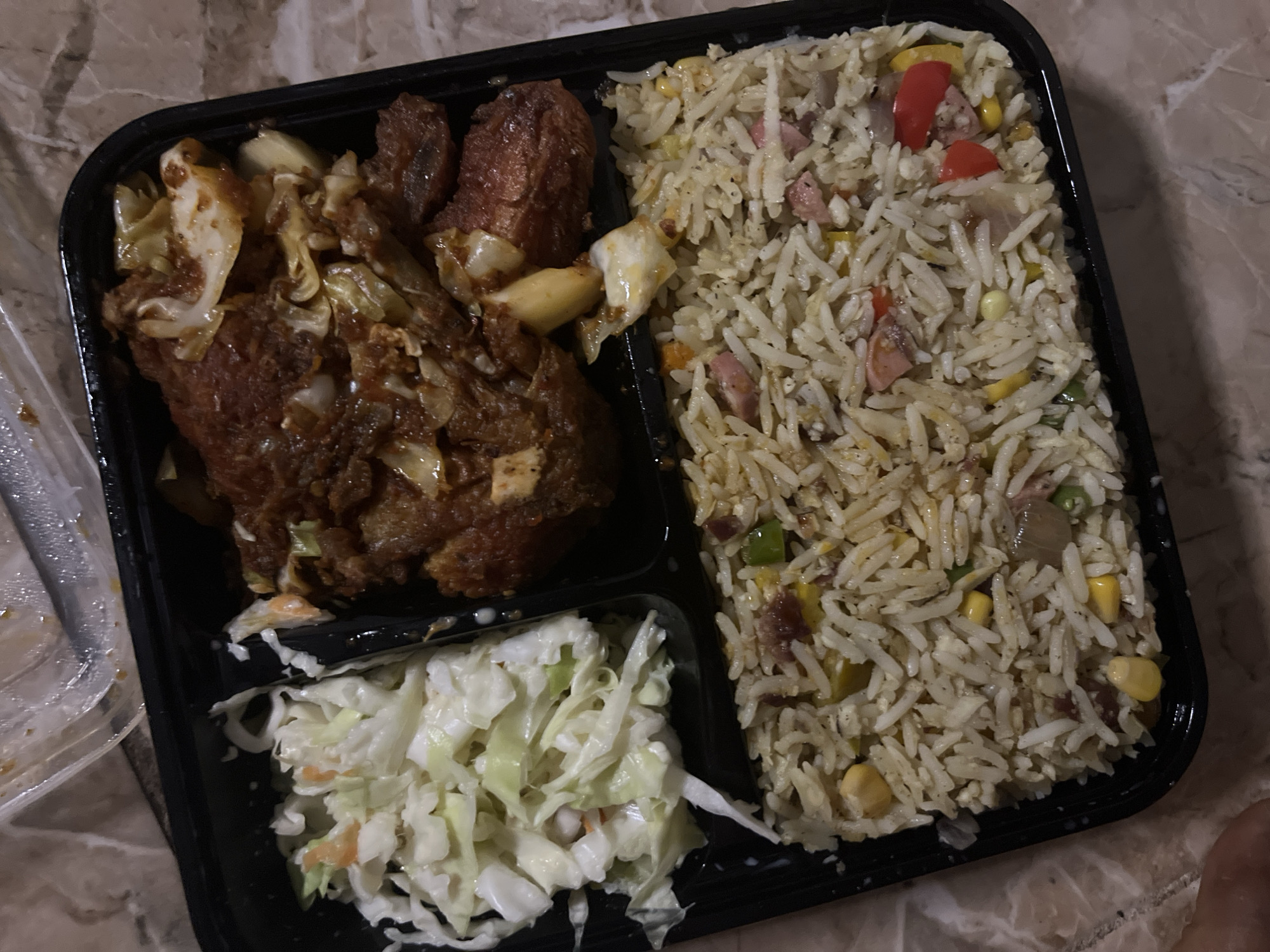 The image shows a black takeout container with three compartments. The upper left compartment contains fried chicken with what appears to be cabbage. The upper right compartment is filled with fried rice containing small pieces of meat, corn, peas, and red peppers. The lower left compartment contains coleslaw. The food is on a surface that resembles a marble countertop. There is no indication of the city of Jalingo, Nigeria. No people are fully visible.