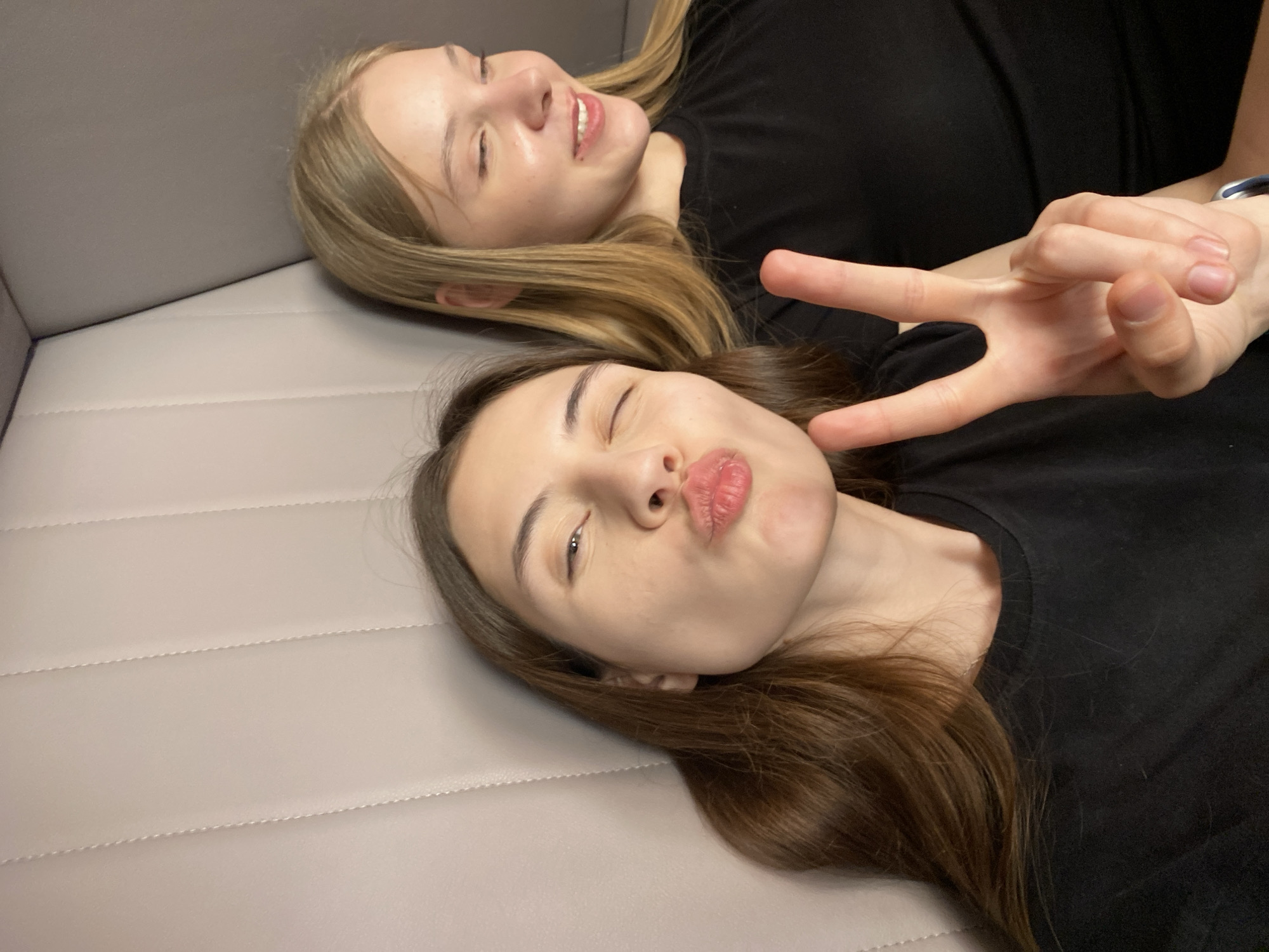 Two young women are lying down next to each other. The woman in the foreground has long brown hair, closed eyes, pursed lips and is holding up the peace sign with her right hand. The woman behind her has long blonde hair, closed eyes and is smiling. Both women are wearing black shirts. They are lying on a gray padded surface. There is no indication of the city of Votkinsk, Russia.
