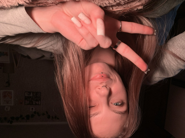An indoor scene in the city of Фоки, Russia, depicts a young, light-skinned female subject captured from the chest up, facing the viewer. She has long, straight, light brown hair flowing over her shoulders. Her left eye (viewer's right) is open, revealing a blue iris, while her right eye (viewer's left) is closed or winking. She exhibits a subtle smile. Facial piercings include a ring in her left nostril and two labret studs on her lower lip. She is wearing a light gray long-sleeved top. Her left hand (viewer's right) is raised, displaying a "V" or peace sign with her index and middle fingers extended. This hand bears two rings: one dark band on the index finger and one silver band on the middle finger. Her fingernails are long, squared, and white. Her left arm rests on a textured, reddish-brown surface, likely a blanket or bedding. The background is dimly lit and out of focus, showing dark vine-like decorations along the upper wall, and multiple framed items or decorations on the wall behind her. The overall lighting has a reddish-pink hue.