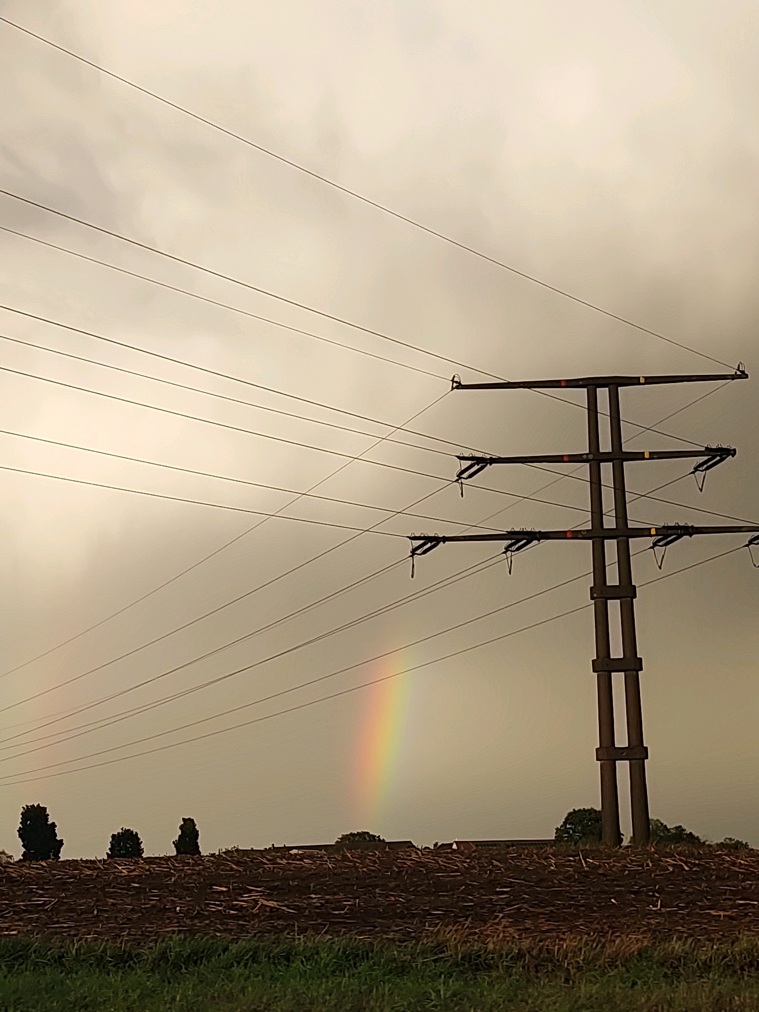 The image shows a power line tower in a field under a cloudy sky. A faint rainbow is visible on the left side of the image. The foreground consists of a pile of wood debris and some grass. In the background, there are several trees and some buildings. The power line tower is tall and dark brown, with multiple crossbars supporting the electrical wires. The electrical wires stretch across the frame, creating a linear pattern against the sky. The sky is overcast with grey clouds, suggesting it may have rained recently. The overall scene appears to be a rural landscape in Lichtenau, Germany.