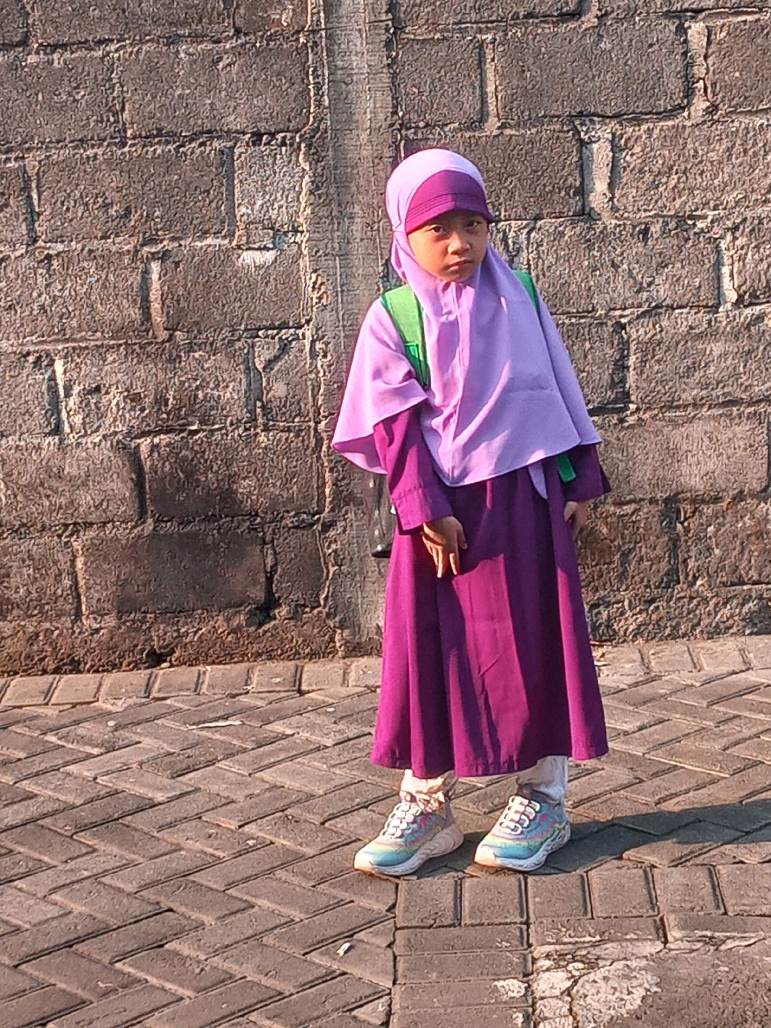 A young girl wearing a purple hijab and dress stands on a brick pathway in front of a brick wall. She has a green backpack on her back and is wearing colorful sneakers. The scene appears to be outdoors on a sunny day, as indicated by the shadows cast on the pathway. The brick wall is rough and uneven, suggesting it might be an older structure. The pathway is laid with rectangular bricks arranged in a herringbone pattern. The overall environment suggests a public space, possibly a street or a park in the Banguntapan sub-district of Bantul Regency, Indonesia, given the commonality of brick structures and pathways in the region.