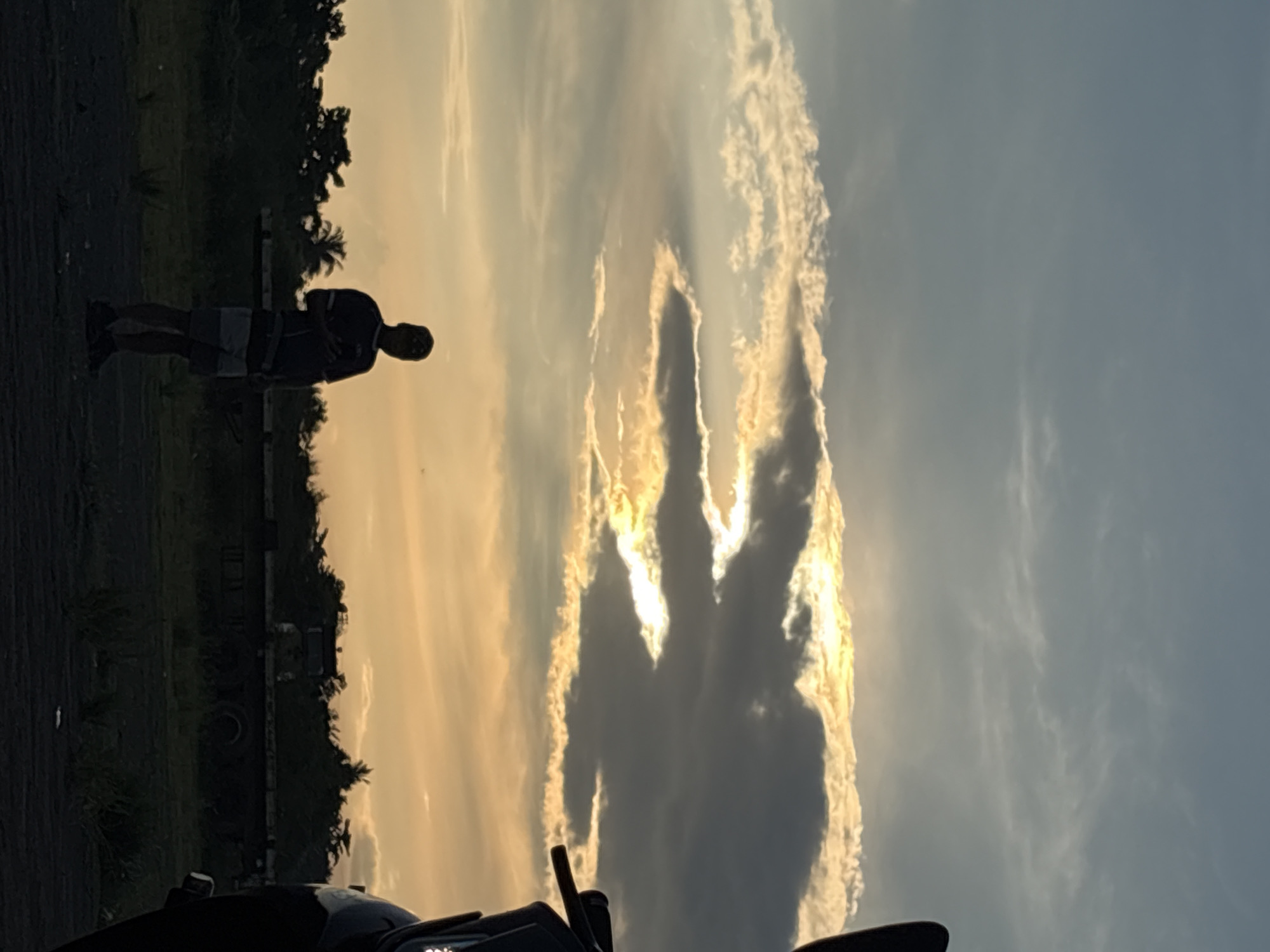 In the city of Davao City, Philippines, at sunset, a lone individual stands silhouetted against a dramatic sky. The sun, partially obscured by dark clouds, casts a golden glow through openings in the clouds. The person, wearing a dark t-shirt and shorts, holds a smartphone in their right hand. The foreground is a patch of dark ground, with sparse vegetation. In the background, a line of trees and a flatbed trailer are visible. To the right, the silhouette of a motorcycle is partially visible. The overall scene is one of a peaceful evening, with the natural beauty of the sky taking center stage.