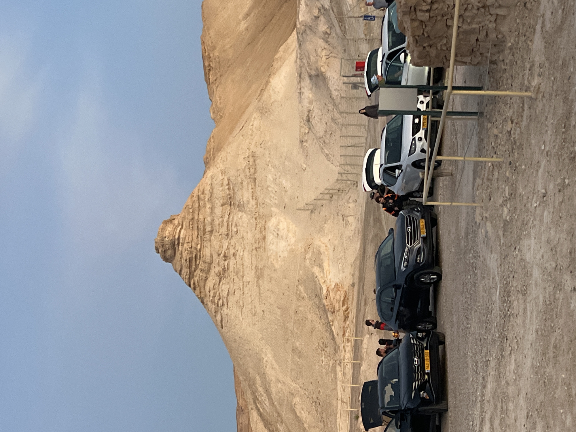 The image depicts a parking lot adjacent to a large, steep, light-brown rock formation under a pale blue sky, likely in the vicinity of the Tamar Regional Council, Israel. Several vehicles are parked in a row on a gravelly surface. They include a white car, two dark-colored SUVs, and another white vehicle with a red light on top, possibly an emergency vehicle. People appear to be standing near the vehicles, though their actions and interactions are unclear due to the distance. A low fence runs along the edge of the parking area, separating it from the rock formation.