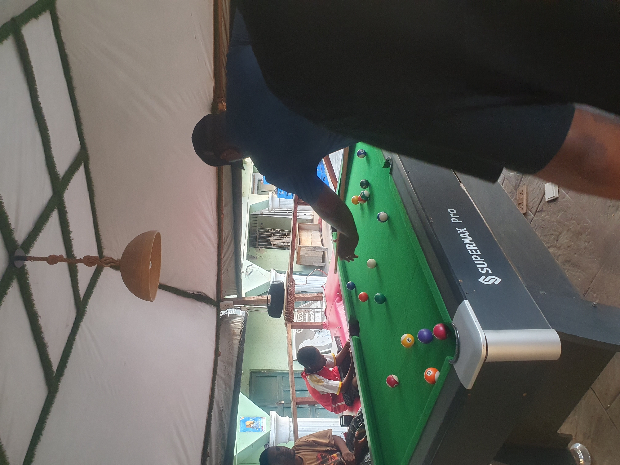 In Lokoja, Nigeria, a group of individuals are playing a game of billiards on a "Supermax Pro" pool table. The billiard table is located within a structure with a white ceiling supported by a frame adorned with green faux grass. A light fixture hangs from the center of the ceiling. One man is taking a shot, while others observe. The setting appears to be a semi-outdoor or covered area, with glimpses of buildings visible in the background.