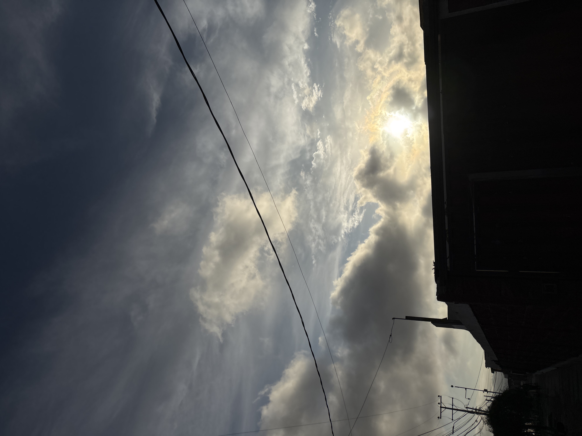 A low-angle, tilted photograph captures a cloudy sky in Arica, Chile. The sun is visible, obscured by clouds, casting a bright glow. Power lines diagonally cross the frame. The silhouette of a building occupies the right side of the image. In the bottom right, power poles and vegetation are visible. The sky ranges from dark gray to light gray with scattered white clouds.