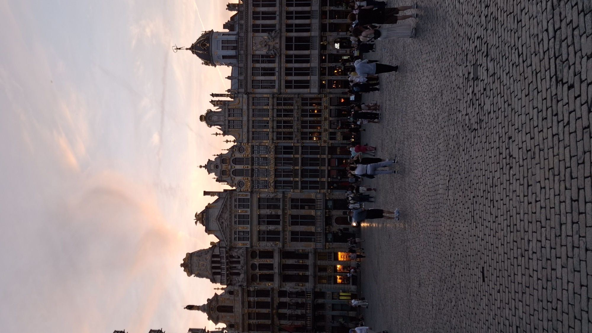 In an open, cobblestone plaza in the city of Unknown, Unknown, a row of ornate, multi-story buildings with gold detailing lines the background. The architecture suggests a historic European city square, with elements of Baroque or Gothic styles. A soft, pastel-colored sky with faint clouds dominates the upper portion of the frame, indicating either sunrise or sunset. Several people are scattered across the plaza, engaged in walking and standing. Some are captured in mid-stride, while others are stationary, observing the surroundings. The lighting is dim, consistent with twilight, casting subtle shadows on the cobblestones. The overall atmosphere is serene and picturesque, evoking a sense of calm amidst urban grandeur.