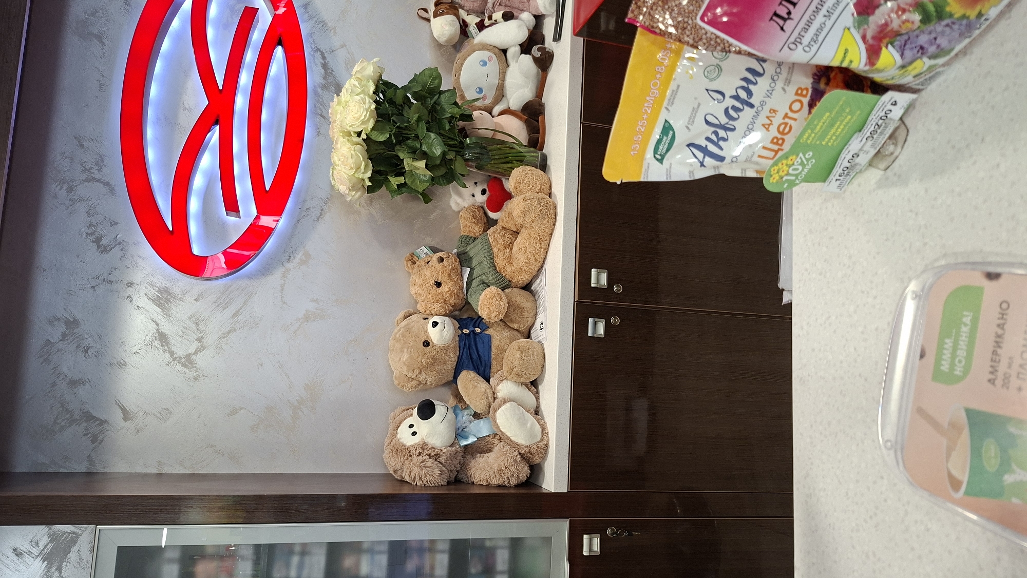 The image depicts an indoor scene, likely a retail environment in Nytva, Russia, given the context. In the foreground, a white countertop is visible, with various products displayed. To the right, a yellow bag with Russian text and graphics of flowers is prominent, possibly indicating a fertilizer or plant-related product. Next to it, a bag of what appears to be grain or seeds is partially visible. A clear plastic display with text in Russian, including "АМЕРИКАНО", suggests a beverage offering.
Further back, on a dark brown cabinet or shelf, a collection of stuffed animals is arranged. These include several teddy bears of varying sizes, a dog-like plush toy, and other assorted characters. Behind the stuffed animals, a vase filled with a bouquet of white roses is placed, with a distinctive red logo featuring a stylized letter 'R' illuminated with white LED lights mounted on a textured, light-colored wall. A glass-fronted cabinet is visible to the far left. The overall impression is of a well-lit retail space with a mix of plush toys, floral arrangements, and consumer goods.