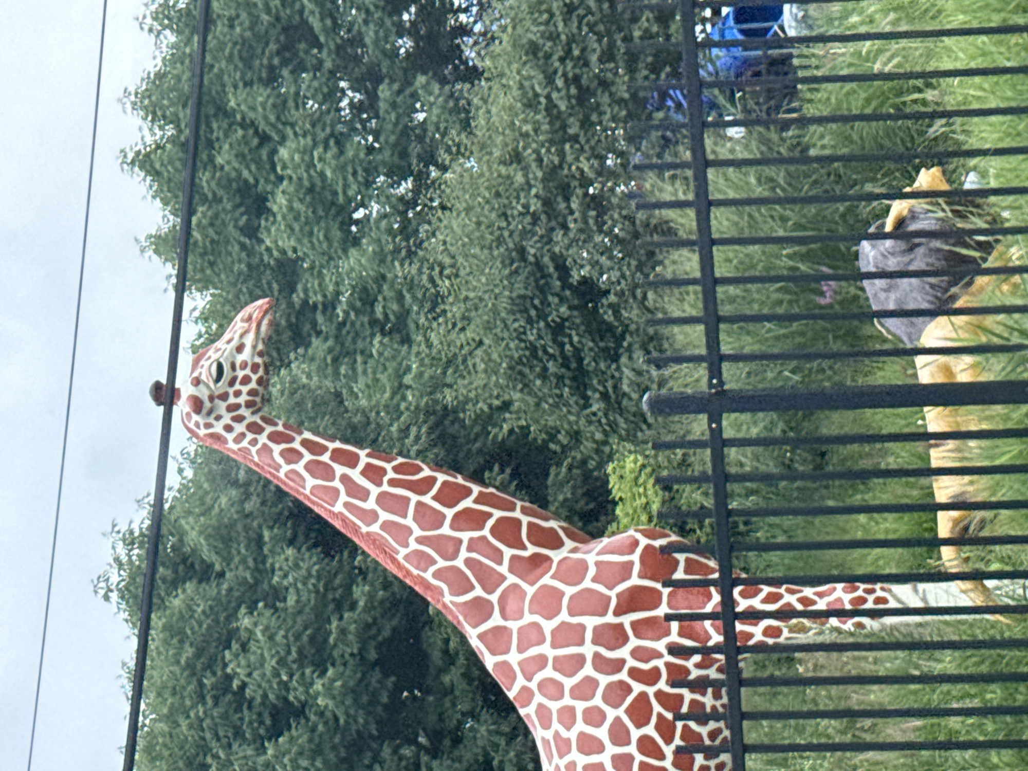 The image shows a statue of a giraffe in front of a black metal fence. The giraffe is reddish-brown with white spots. Behind the fence, a statue of a lion is visible, lying down. Green trees and foliage are in the background, with a cloudy sky above. The scene appears to be outdoors, possibly in a park or zoo. The location is Skokie, United States of America.