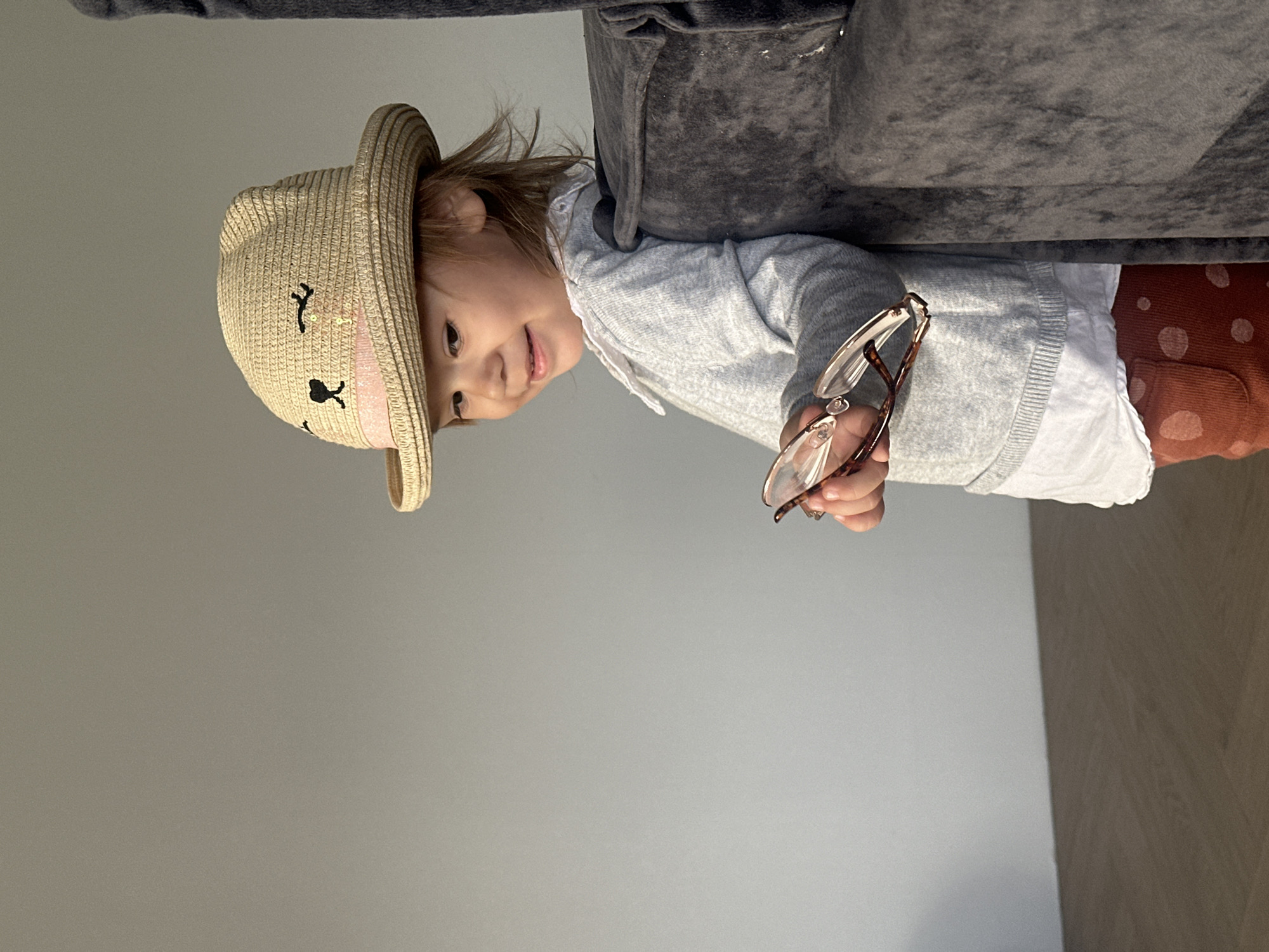 A young child is depicted in an indoor setting in Kharnavy, Russia, with the image rotated 90 degrees clockwise. The child, likely a toddler or preschooler, is wearing a light-colored straw hat with an embroidered cat face design, featuring black eyes, nose, whiskers, and pink inner ears. Light brown hair is visible beneath the hat.
The child has a gentle, slightly smiling expression and is looking upwards. They are dressed in a light grey long-sleeved hooded top or cardigan over a white collared shirt, and reddish-brown pants with white polka dots. Both hands are holding a pair of light brown eyeglasses.
A dark grey upholstered furniture piece, possibly a sofa or armchair, is visible behind and to the upper right of the child. The background is a plain, light grey wall, with a section of a light-colored wooden floor visible in the lower right. The lighting is bright and even, indicating an indoor daytime environment. The child appears to be engaged in playful activity, possibly trying on the hat and glasses. No text is visible in the image.