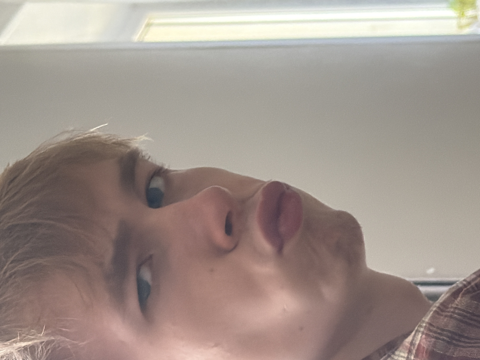This is a close-up, head-and-shoulders portrait of a young person with light brown hair, wearing a plaid shirt. The person is looking towards the camera with their lips pursed in a puckering expression. The background is out of focus and appears to be an indoor setting with a window that lets in natural light. There is no discernible indication of the city of Chaikovsky, Russia.