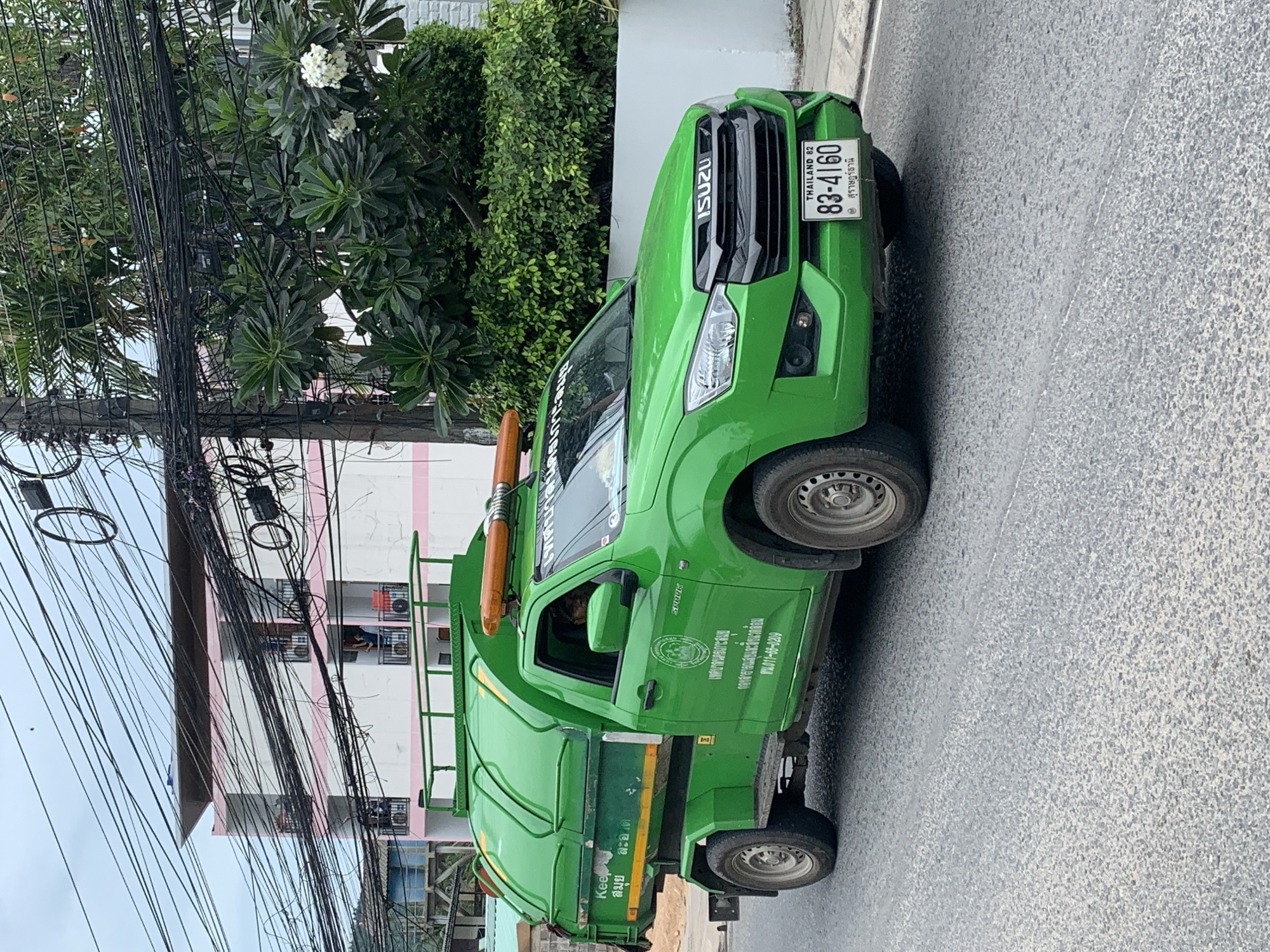 A bright green Isuzu garbage truck is parked on a paved street in the Ko Samui District of Thailand. The truck has a large green bin on the back and an orange flashing light bar on top of the cab. The cab of the truck is green, and the front grille is black with an ISUZU logo. A license plate on the front of the truck reads "THAILAND 82 83-4160". Text in Thai is visible on the side of the truck and on the garbage bin. A person is visible in the driver's seat of the truck. In the background, there is a multi-story building with pink and white trim, a large amount of tangled electrical wires, and lush green trees. The sky is overcast.