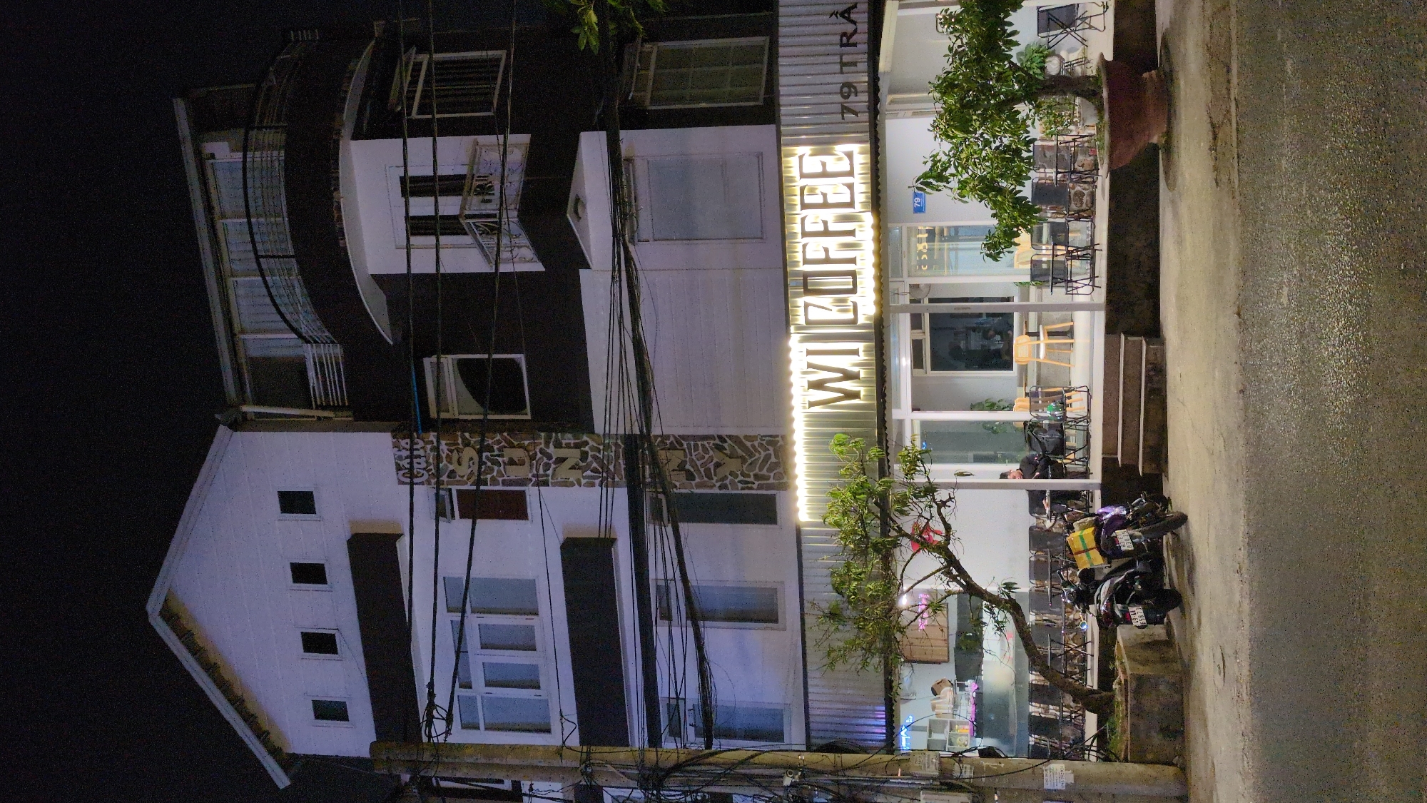 An outdoor scene showing a multi-story building at night. The building is segmented into dark and light sections, with the top floors painted darker. The front of the building is illuminated by the sign "WI COFFEE" and the address "79 TRA." The interior of the coffee shop is visible, with tables and chairs set up. Outside, motorbikes are parked near a small tree. The street appears wet, reflecting the ambient light. The surrounding environment is dark, suggesting nighttime in an urban area. The scene is located in the city of Da Lat, Vietnam.