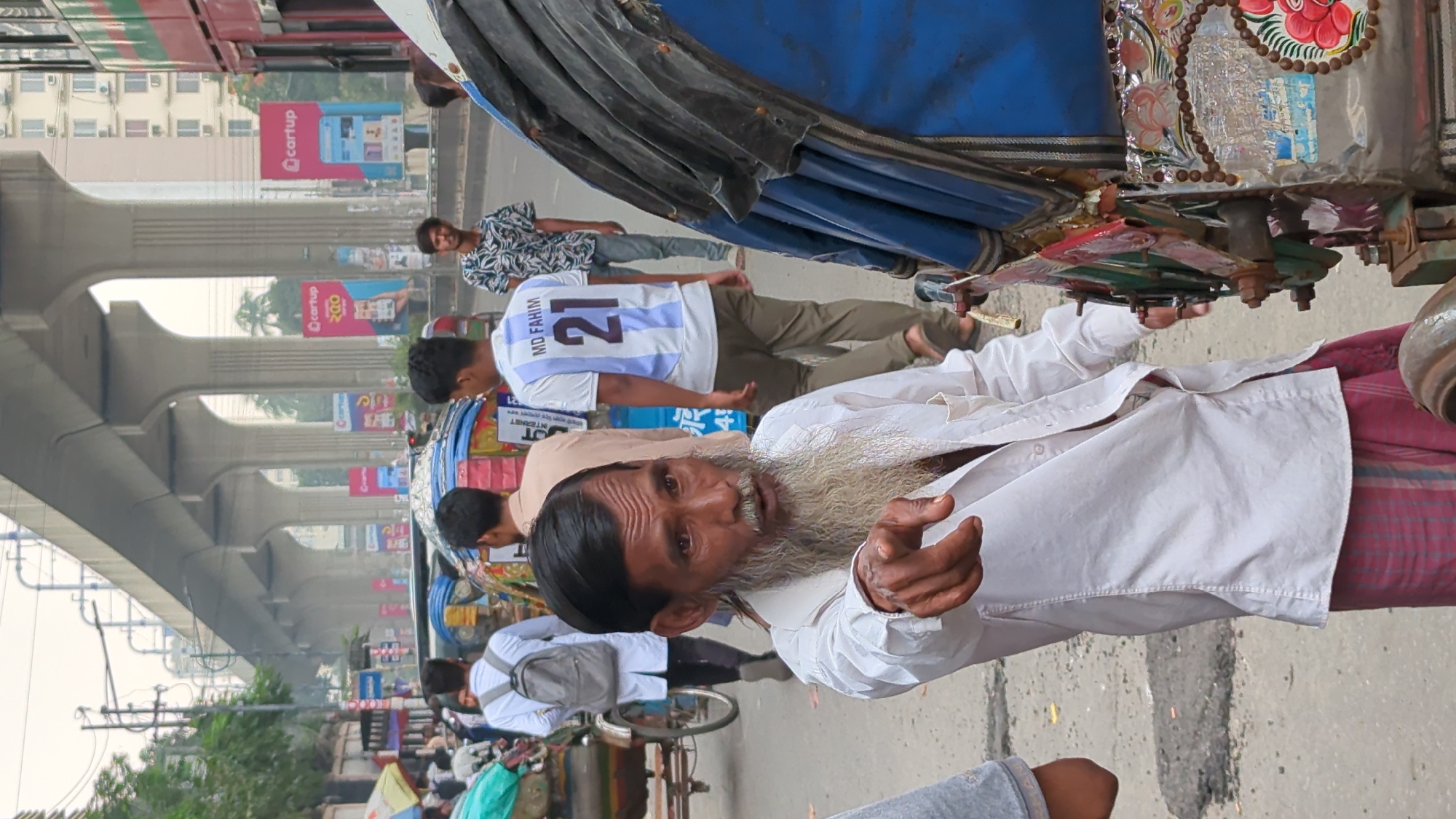 The image depicts a street scene in Dhaka, Bangladesh, under an elevated bridge structure. A man with a long white beard is prominently featured, wearing a white shirt and pointing towards the camera. Behind him, a person wearing a white jersey with the number 21 and the name "MD. FAHIM" is visible. Other individuals are walking on the street, and a rickshaw is partially visible on the right side. The background includes billboards advertising "cartup" and general street clutter.
