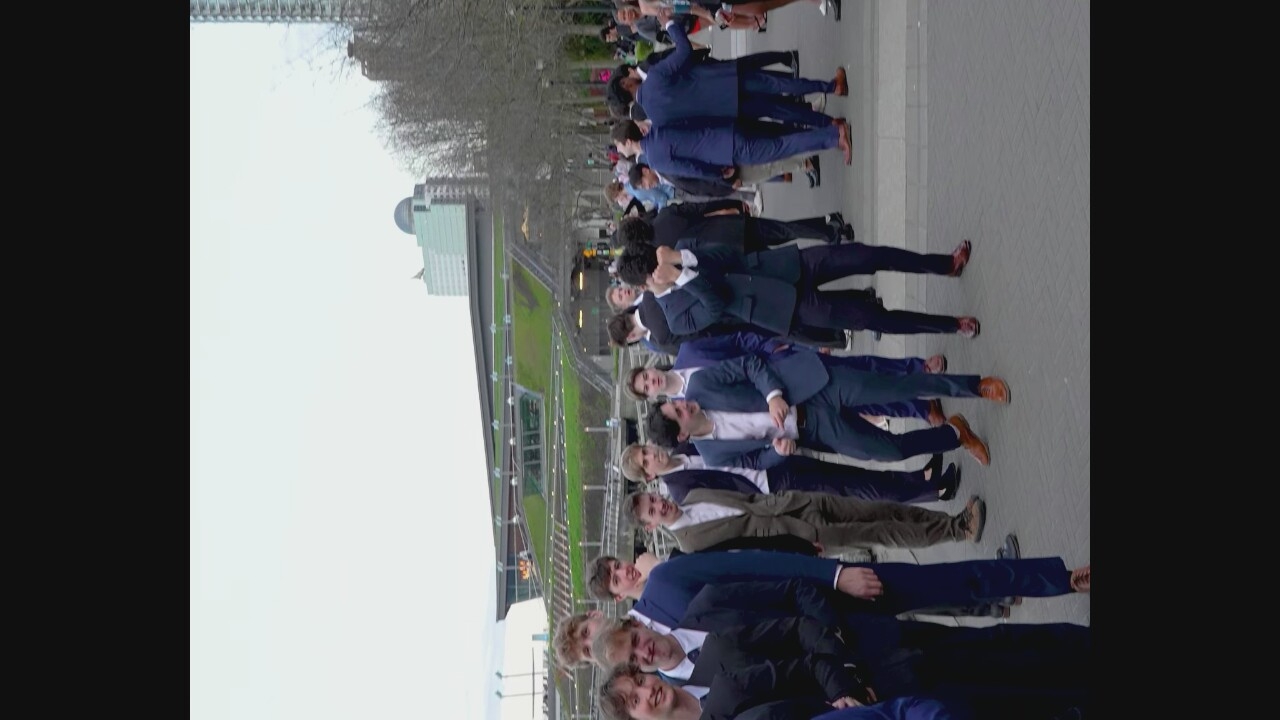 A group of young men in suits stand and interact on a paved outdoor plaza, with a modern building complex visible in the background. The building appears to be a convention center or sports arena with a distinctive green, tiered roof structure, possibly BC Place Stadium in Vancouver, Canada. Several of the men are smiling and looking at each other, while one man in the center is covering his face with his hands, seemingly in distress or amusement. The overall atmosphere suggests a social gathering or formal event. The paving stones are arranged in a basket-weave pattern. The sky is overcast, and trees are visible in the middle distance.