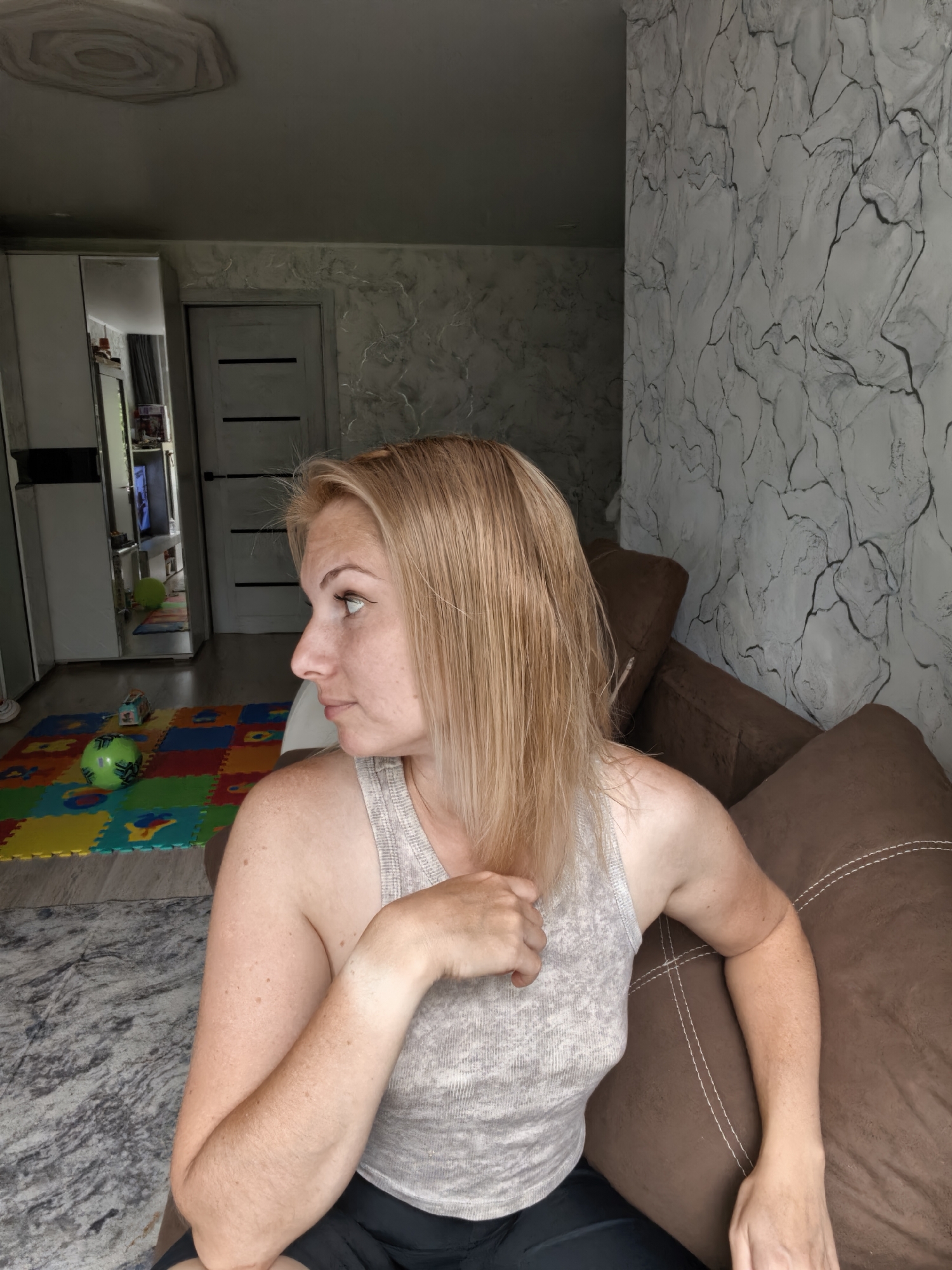 The image shows a medium shot of a woman with blonde hair sitting on a brown sofa. She is wearing a grey tank top and black shorts. Her head is turned to the left, and she is looking away from the camera. The background shows a room with a door, a wall with a textured pattern, and a ceiling with a decorative element. In the foreground, there is a colorful play mat with a green ball on it. The scene appears to be an indoor domestic setting. There is no visual information in the image that would allow for the identification of the location as Sterlitamak, Russia.